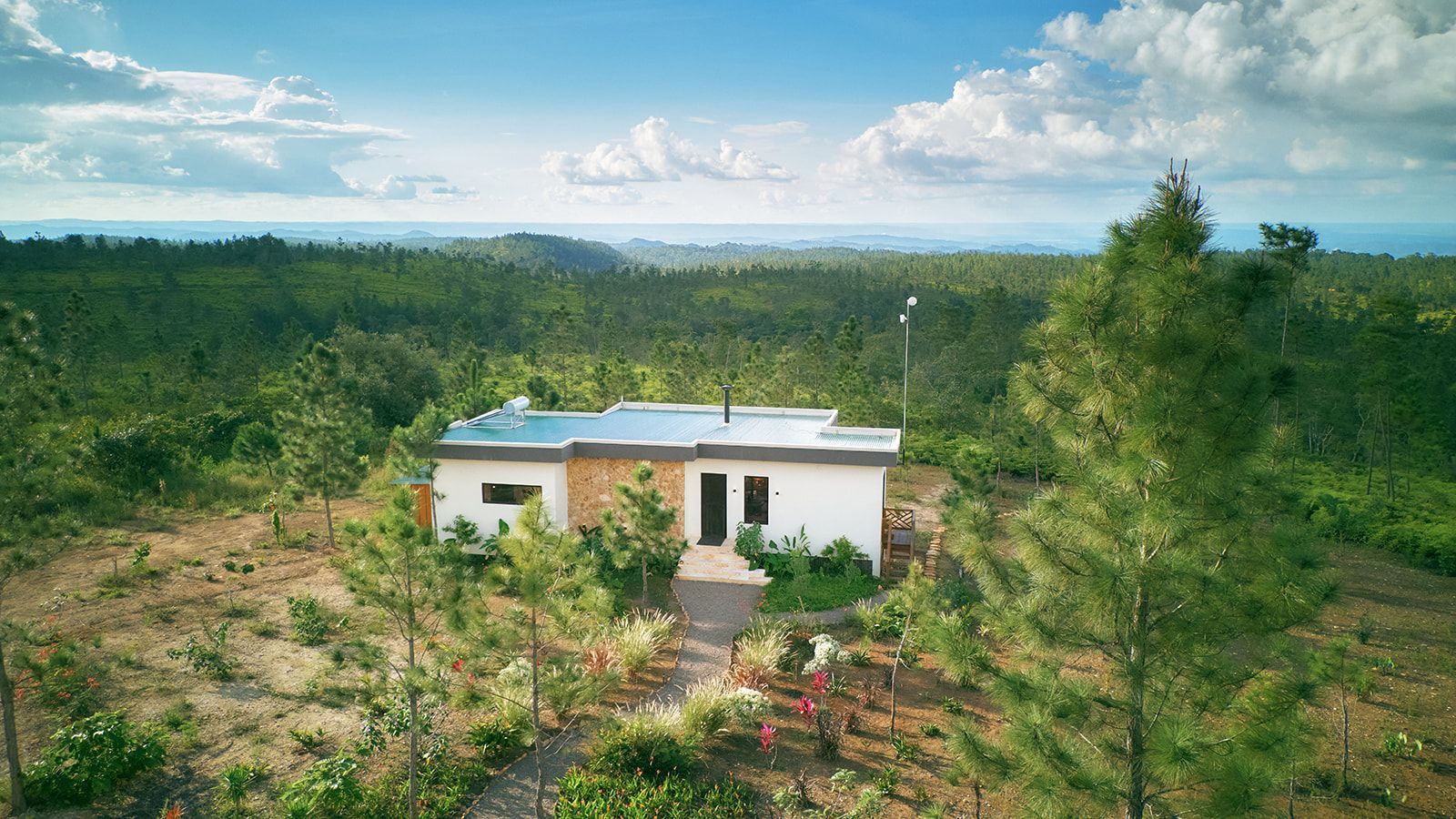 A small white house with a flat roof sits in a lush green forest, under a blue sky with clouds.