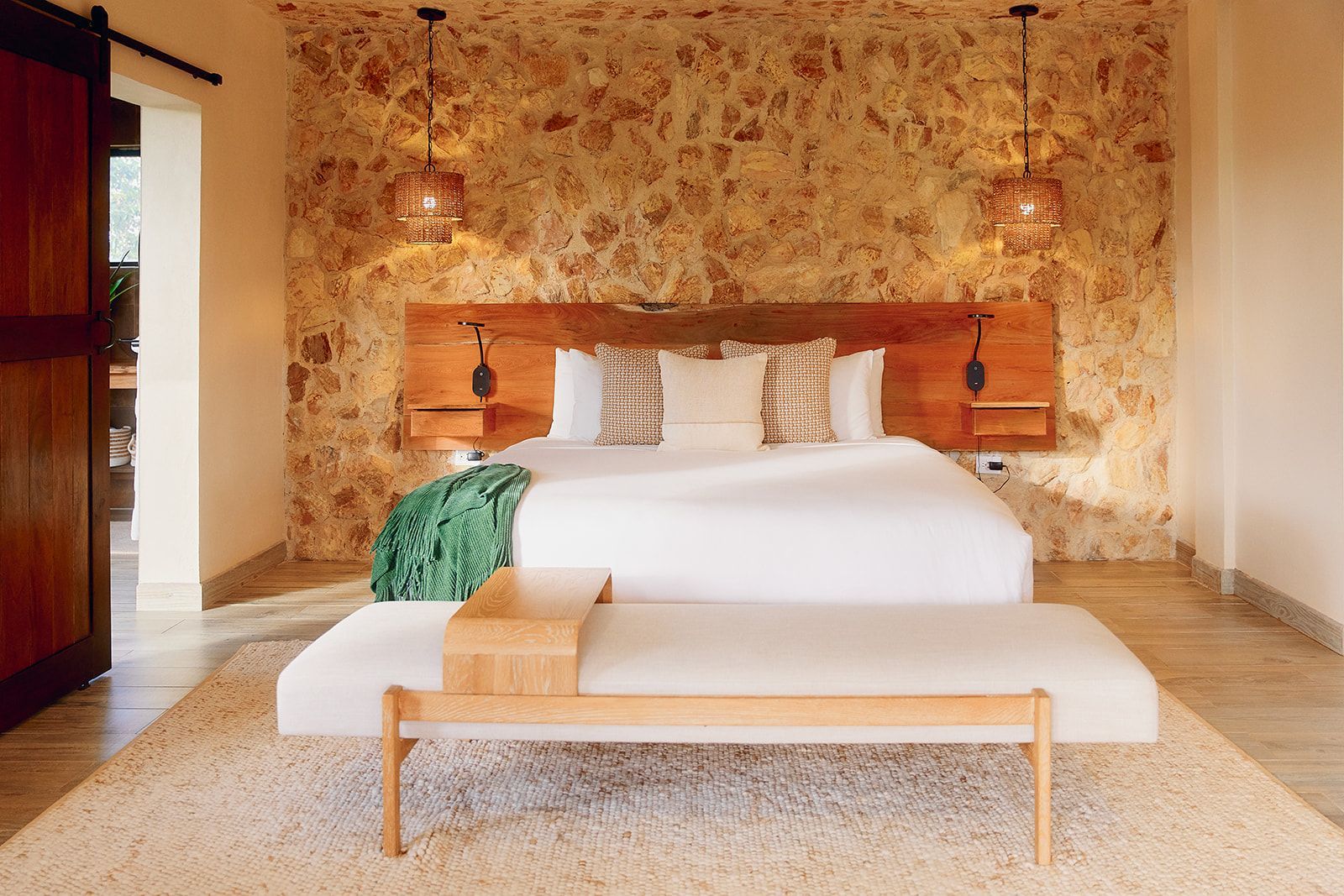 Bedroom with a stone wall, wooden headboard, white bed, bench, and two pendant lights.