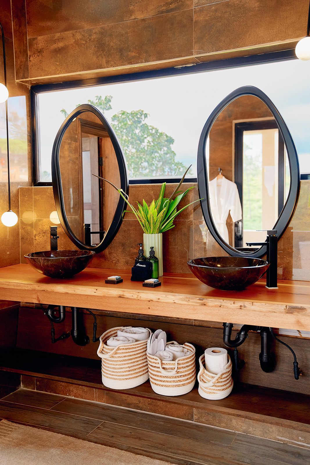 Rustic bathroom with two oval mirrors, brown sinks, and wooden countertop. Baskets and a window view.