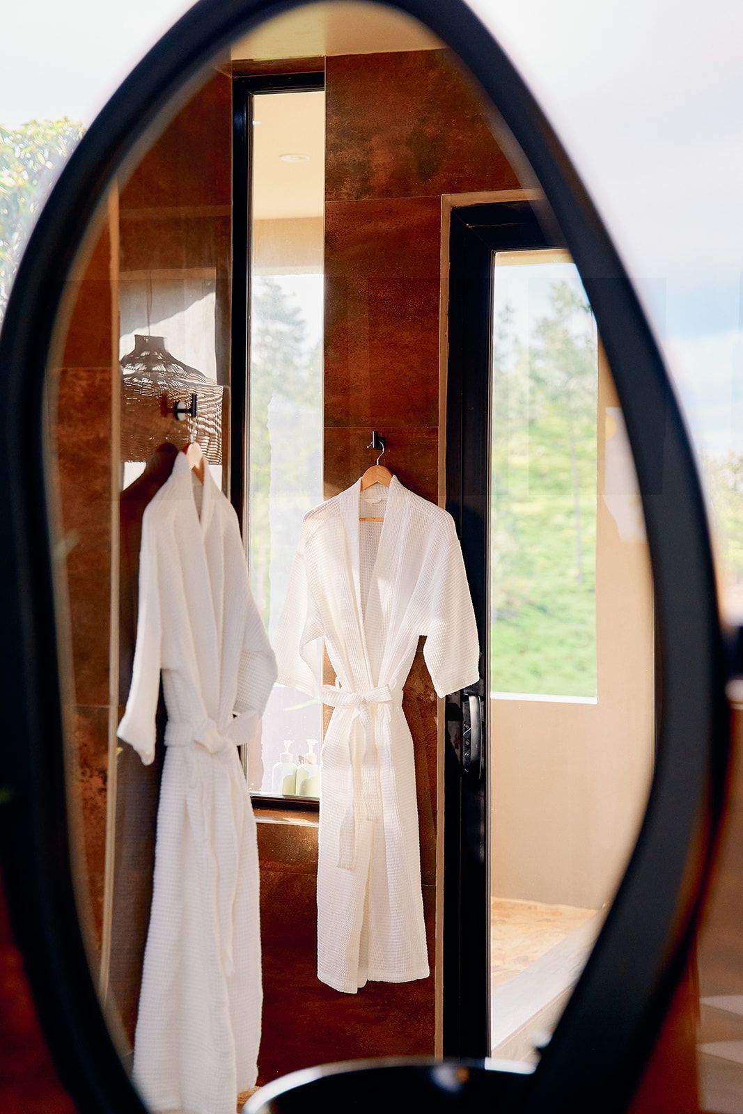 Two white bathrobes hanging in a bathroom, reflected in an oval mirror.