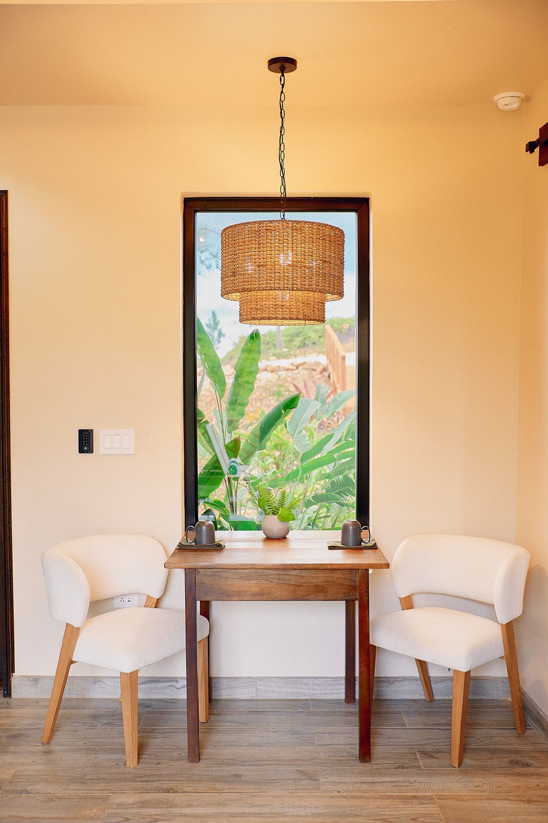 Two white chairs face a small wooden table by a window, with a woven light fixture overhead.