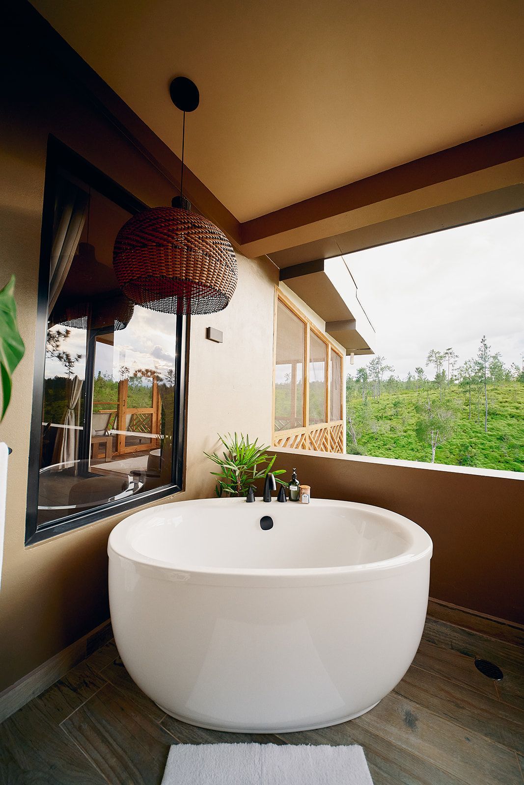 A white, round bathtub on a balcony, with an overhead woven light fixture and a view of green foliage.