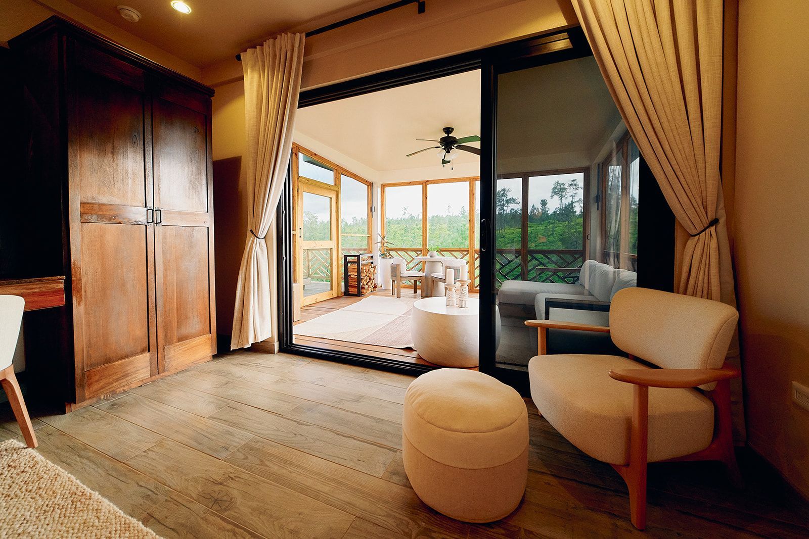 Cozy room with open sliding door to a screened porch. Armchair, ottoman, wooden wardrobe, beige curtains, and nature views.