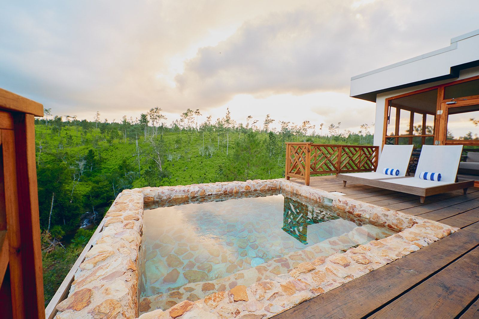Outdoor spa with stone surround, overlooking lush green foliage. Two lounge chairs under a canopy.
