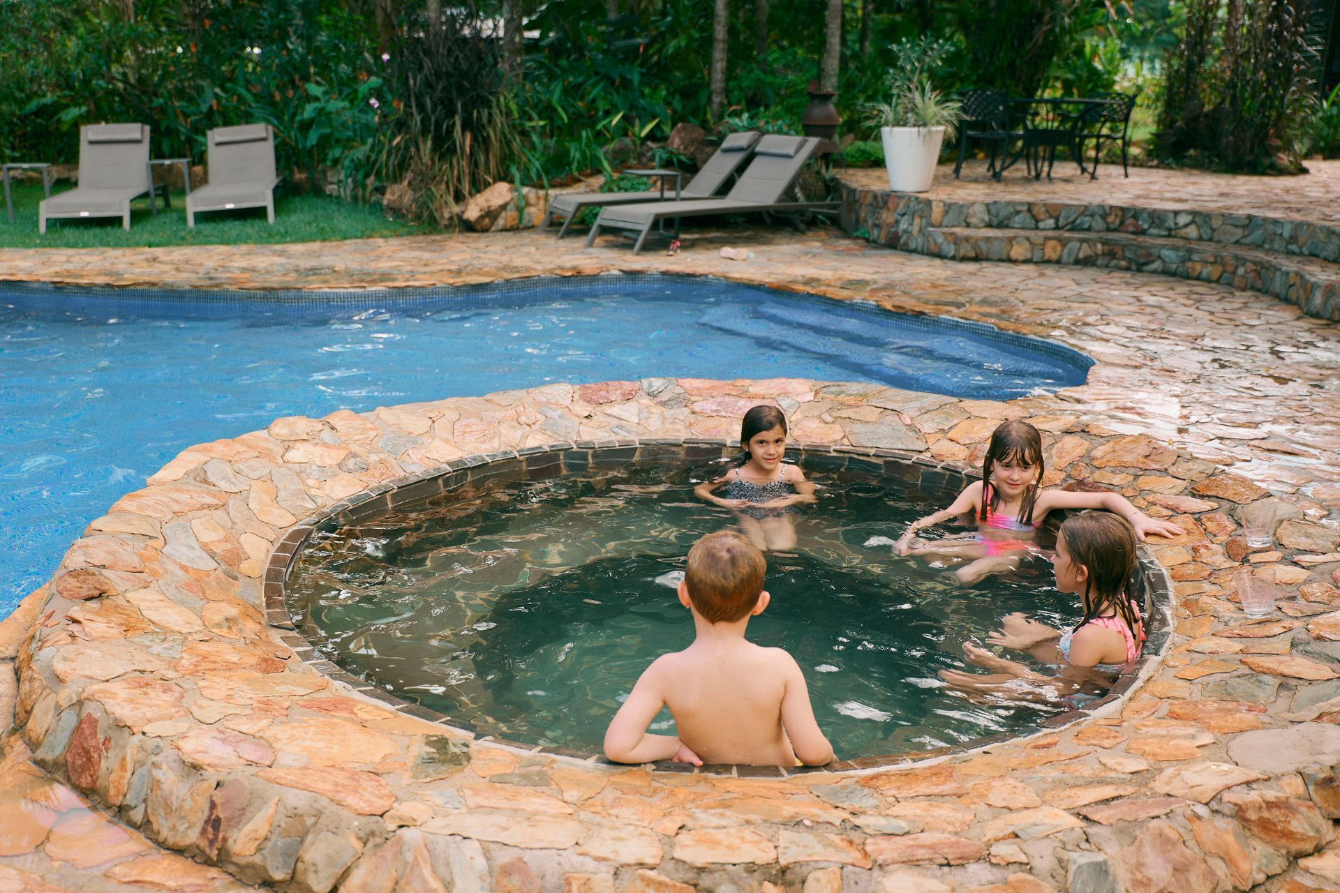 Children in a stone hot tub, near a swimming pool, enjoying themselves outdoors.