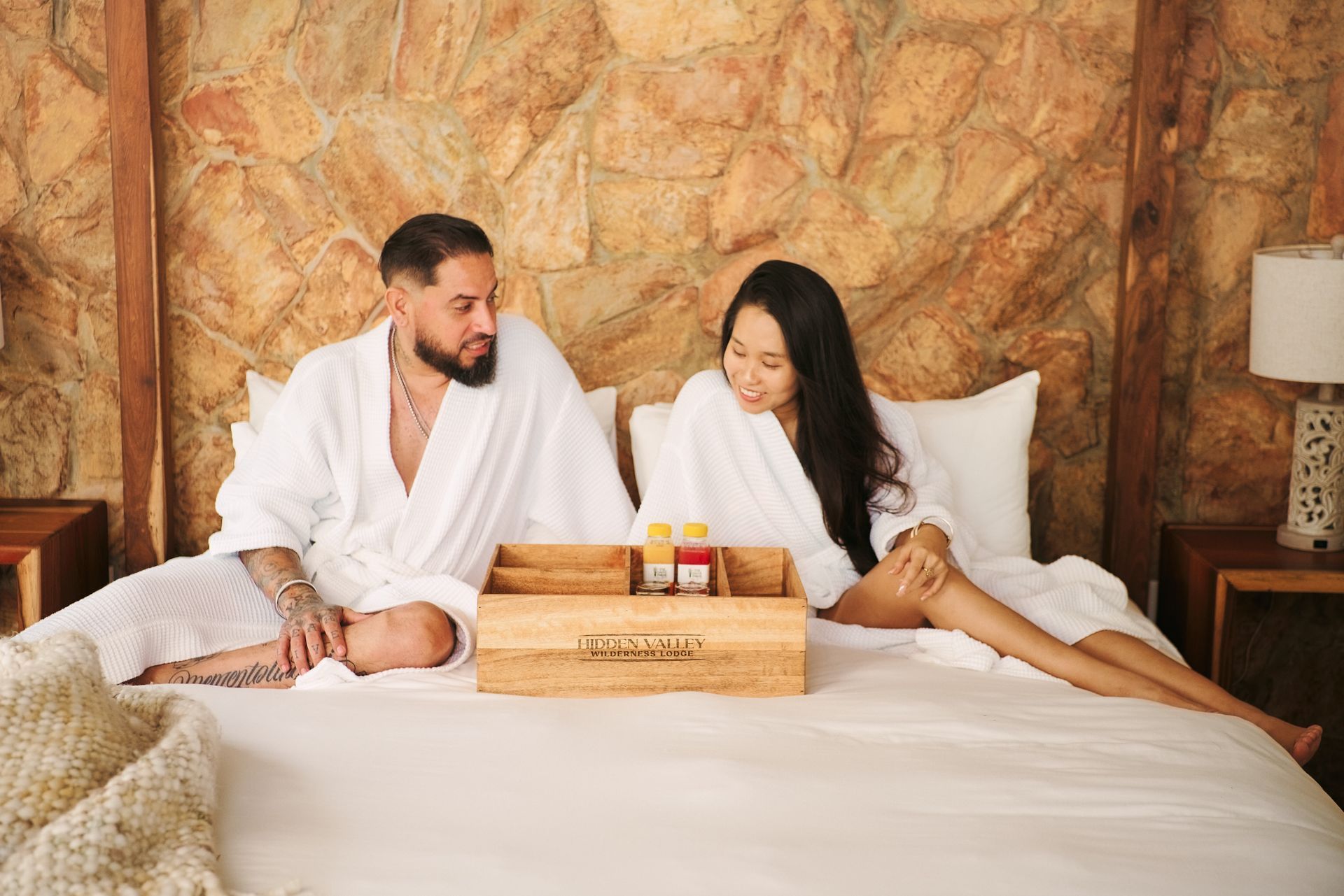 A man and a woman are sitting on a bed with a tray of food.