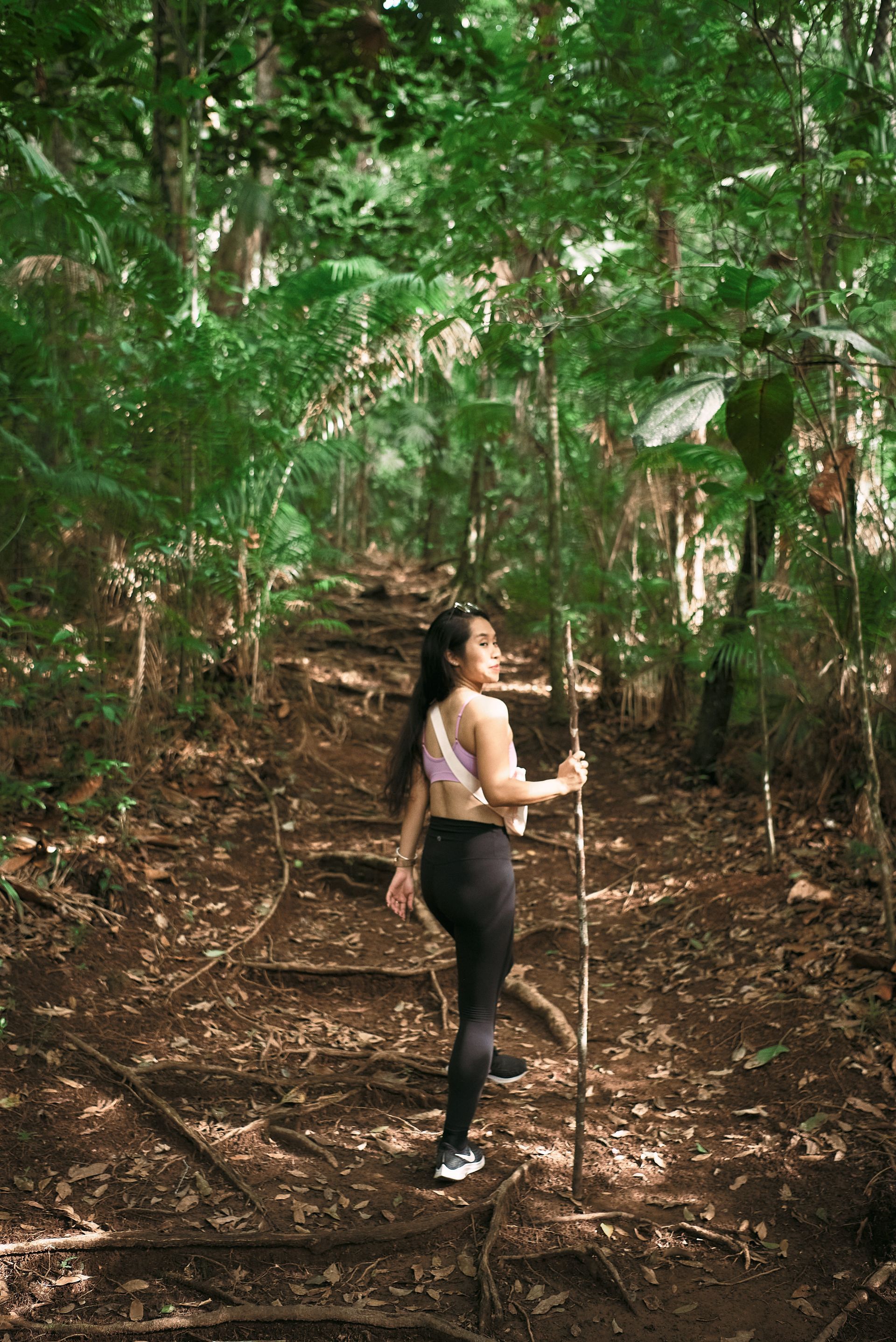 A woman is walking through a lush green forest holding a stick.