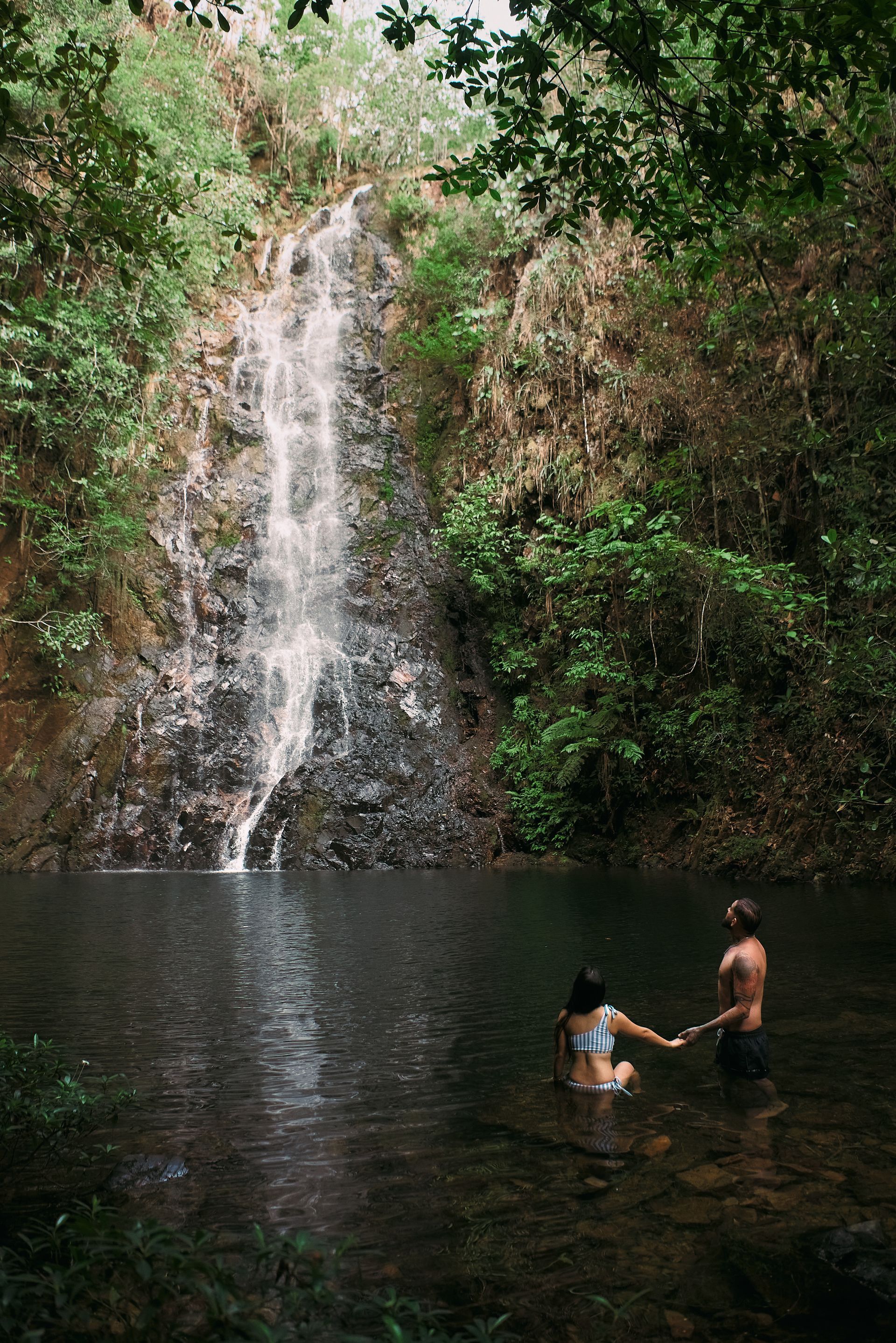 A man and a woman are standing in a pond in front of a waterfall.