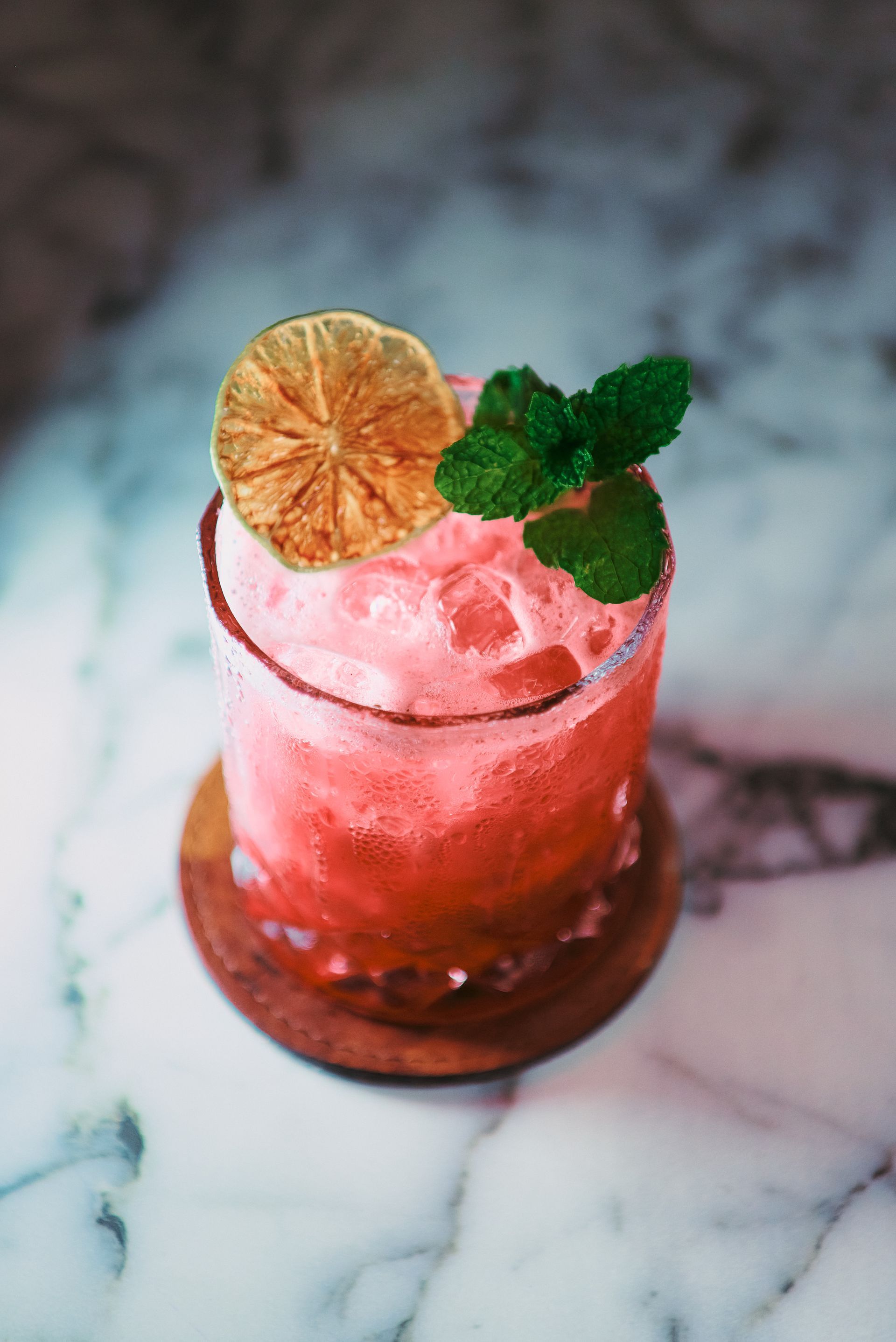 A close up of a pink cocktail with a slice of lemon and mint on a table.