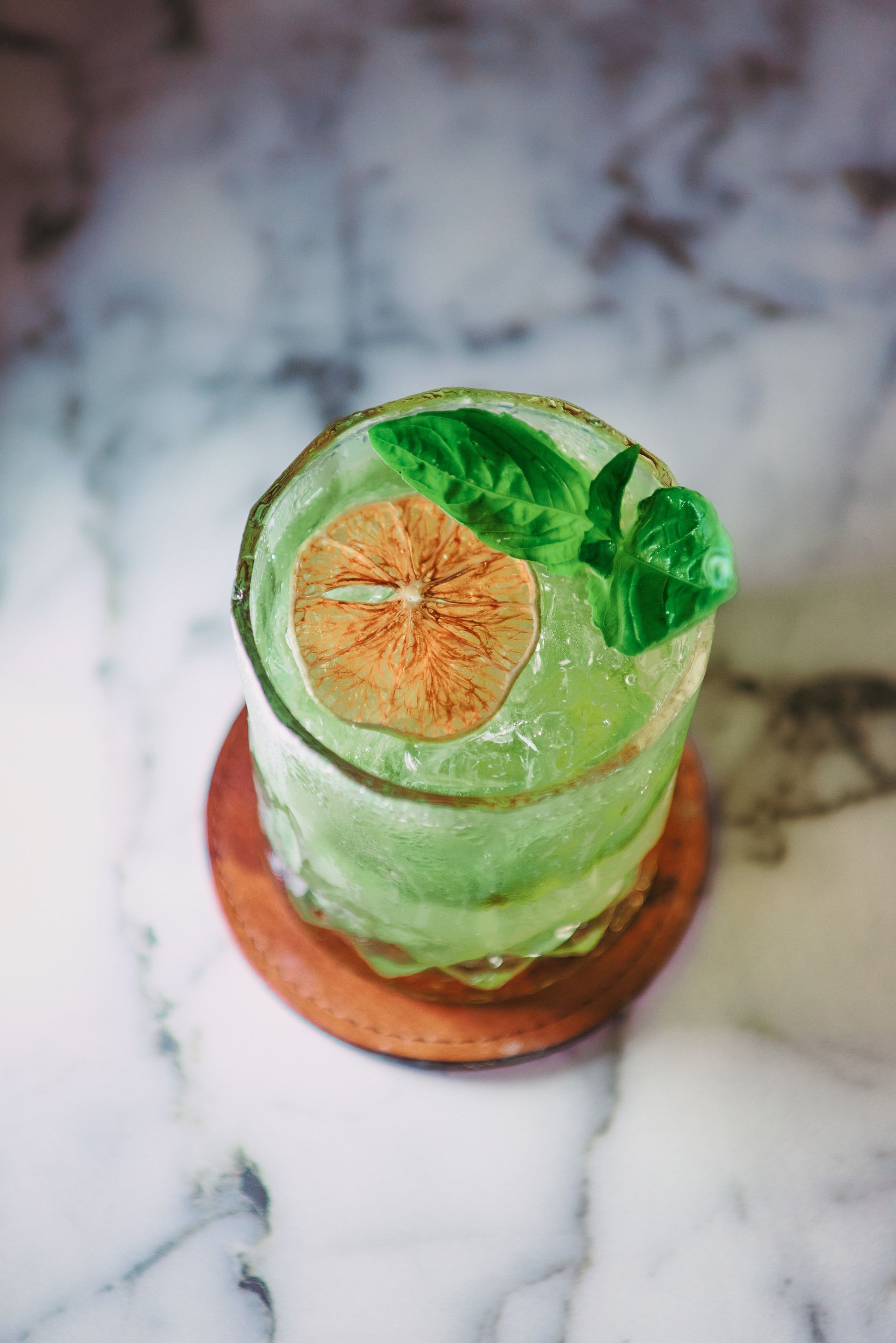 A close up of a green cocktail with a slice of lime on a wooden coaster on a table.