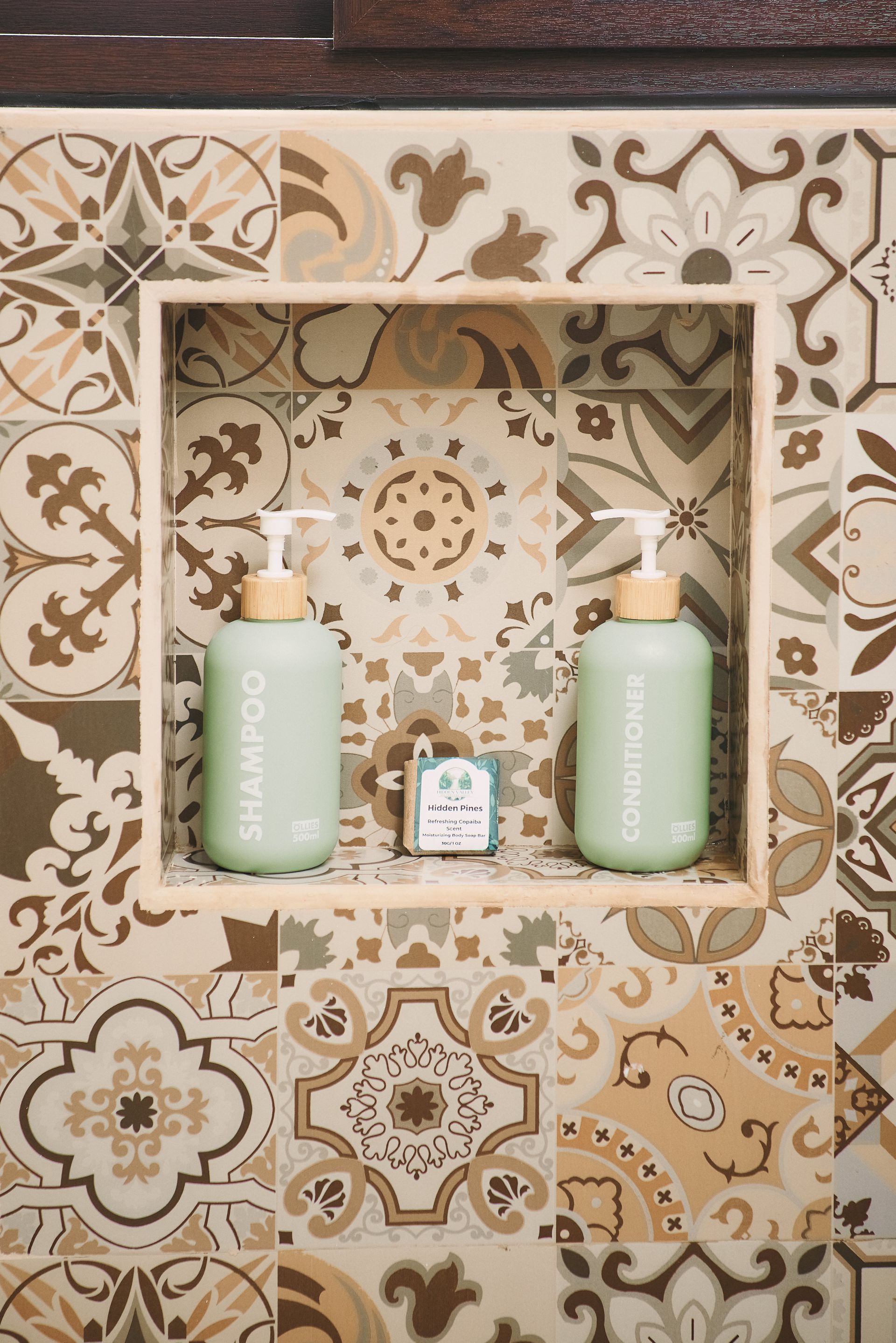 Two bottles of organic shampoo and conditioner are sitting in a niche on a tiled wall.