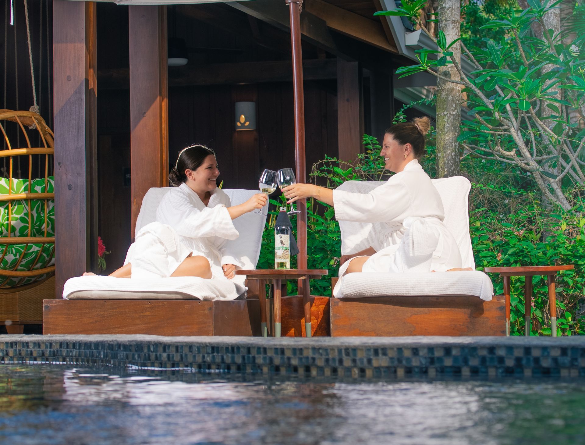 A man and a woman are sitting next to a swimming pool toasting with wine glasses.