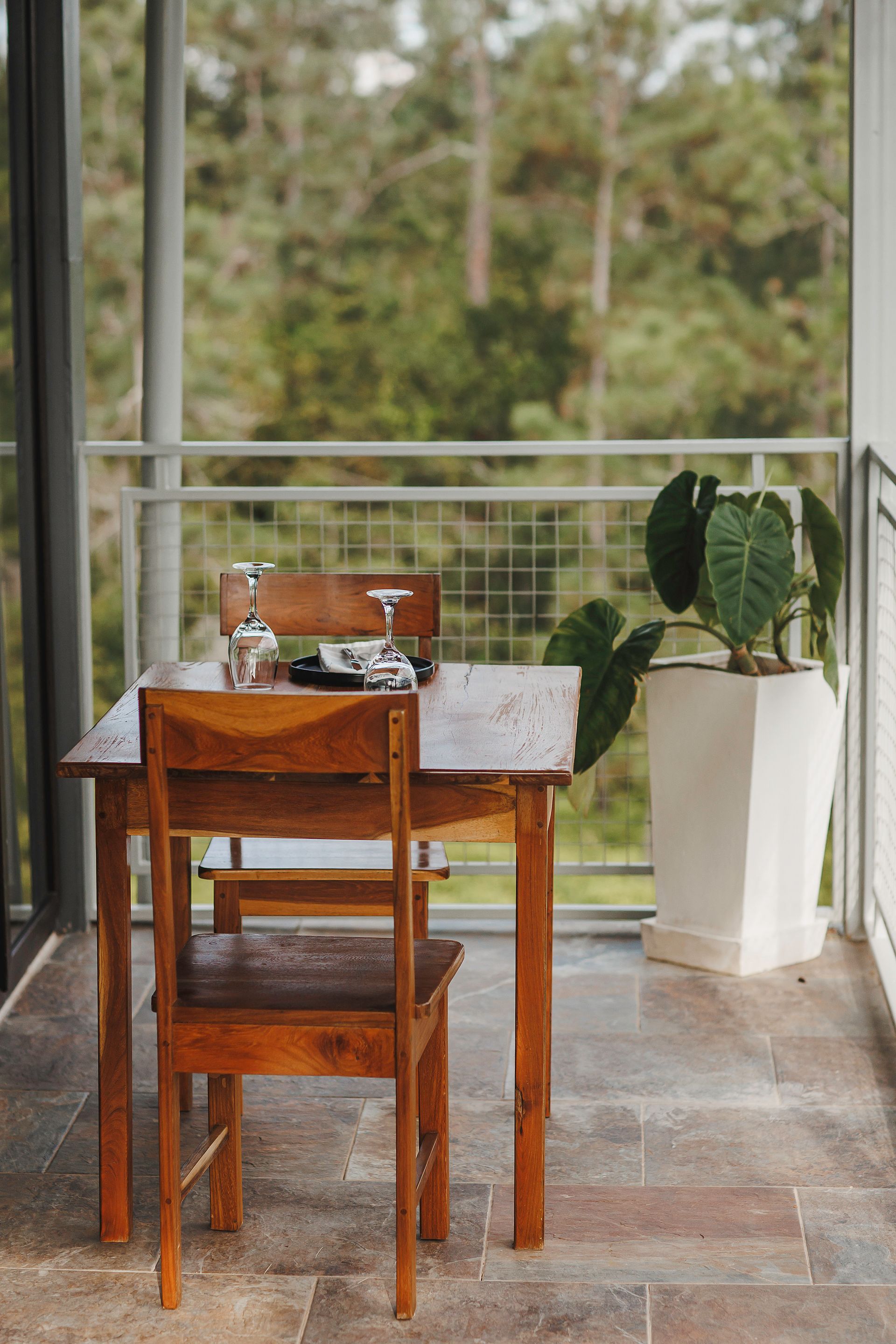 Wooden table and chairs on a balcony overlooking a forest, with potted plant and glasses.