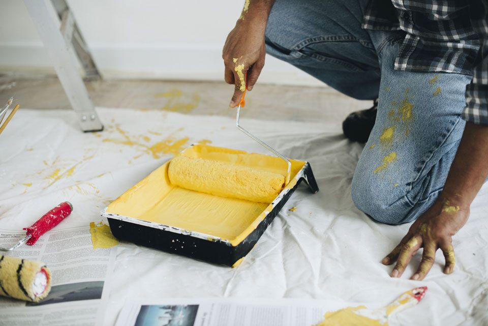 A man is kneeling on the floor painting a wall with a yellow paint roller.