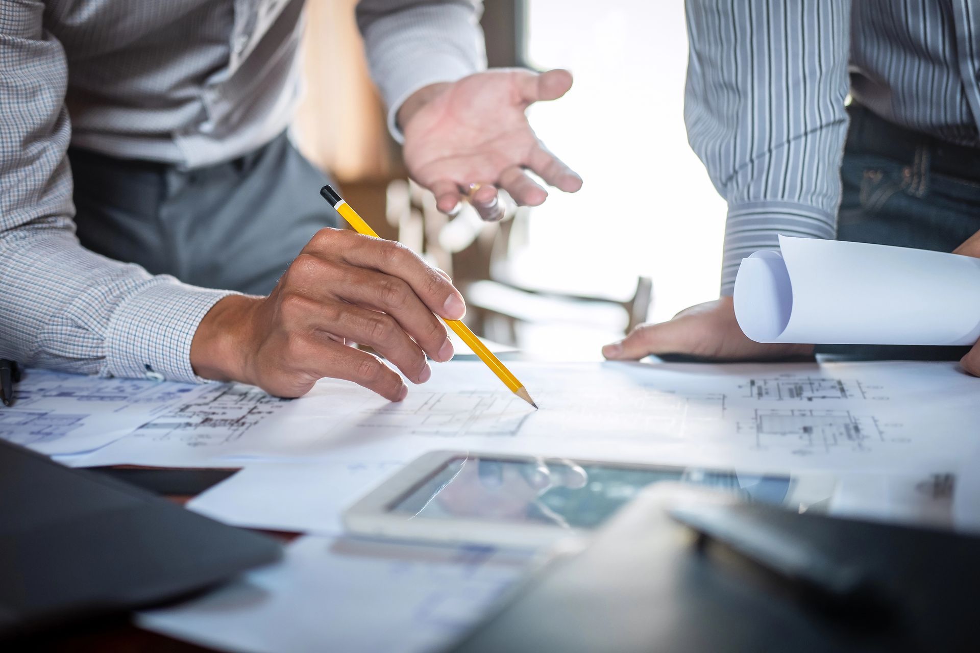 Dos personas discutiendo planos, una señalando con un lápiz, la otra gesticulando con las manos, documentos sobre la mesa.