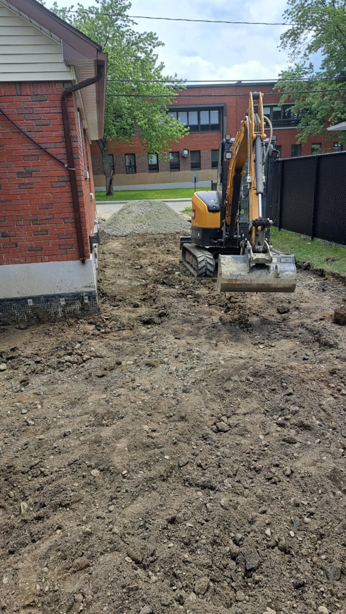 Une petite excavatrice creuse un trou dans la terre devant un bâtiment en briques.