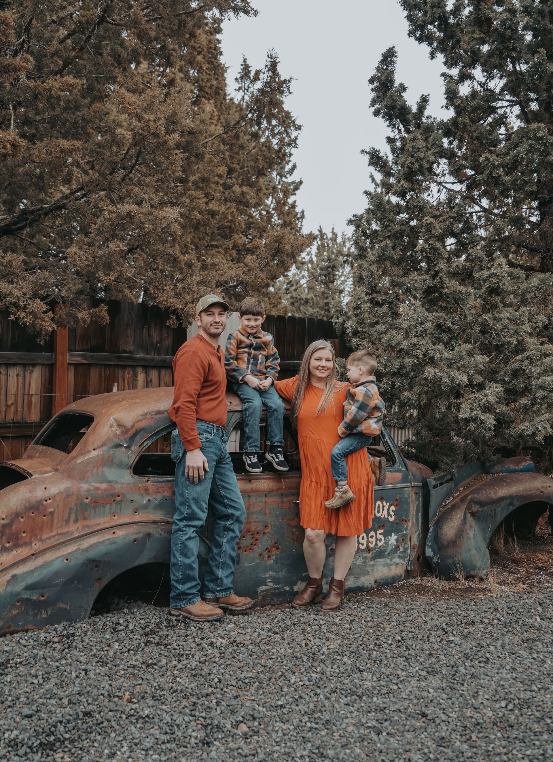 Family posing by a rusted car; man in red shirt, woman in orange dress, and two young boys. Autumn setting.