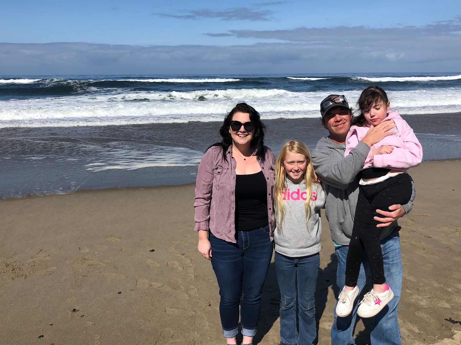 Family of four smiling on a sandy beach. Woman in pink holds a child; other girl and woman stand nearby, ocean waves in the background.