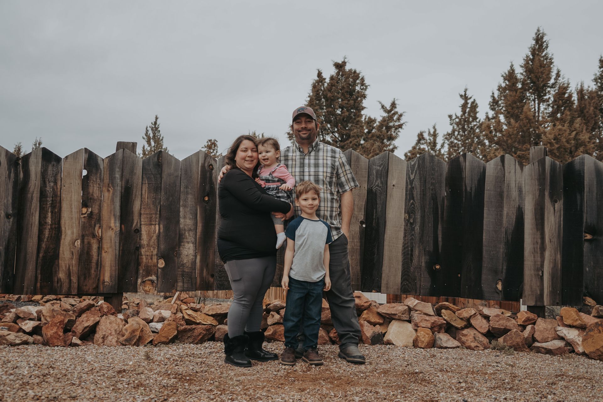 Family of four posing in front of a wooden fence. Overcast sky.