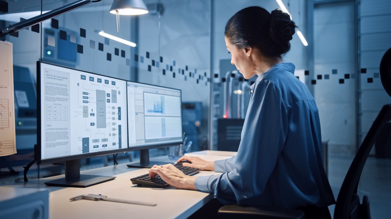 A woman is sitting at a desk in front of three computer monitors.