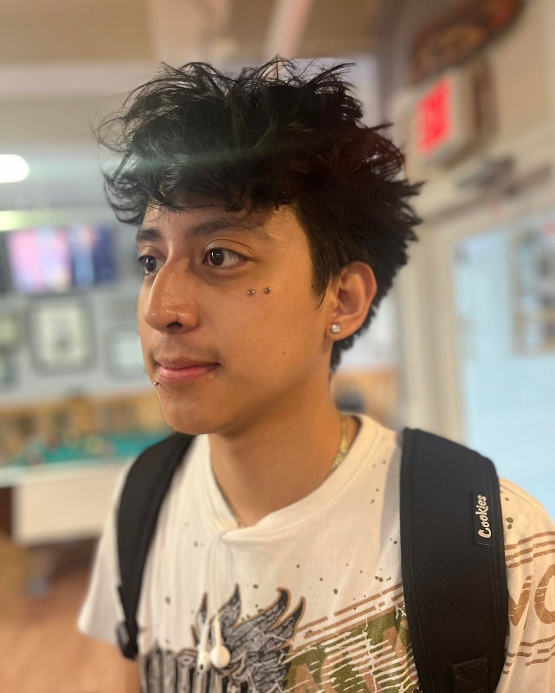 Young person with curly dark hair, wearing a white shirt and backpack, looking off to the side. Inside a building.