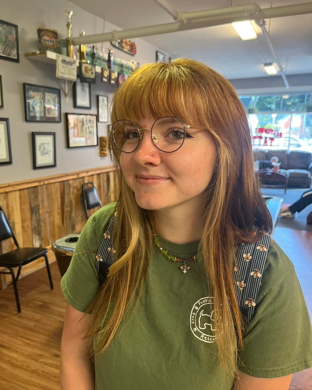 Young person with orange-tinted hair and glasses smiles. Wearing green shirt, in a room with wood paneling and decor.