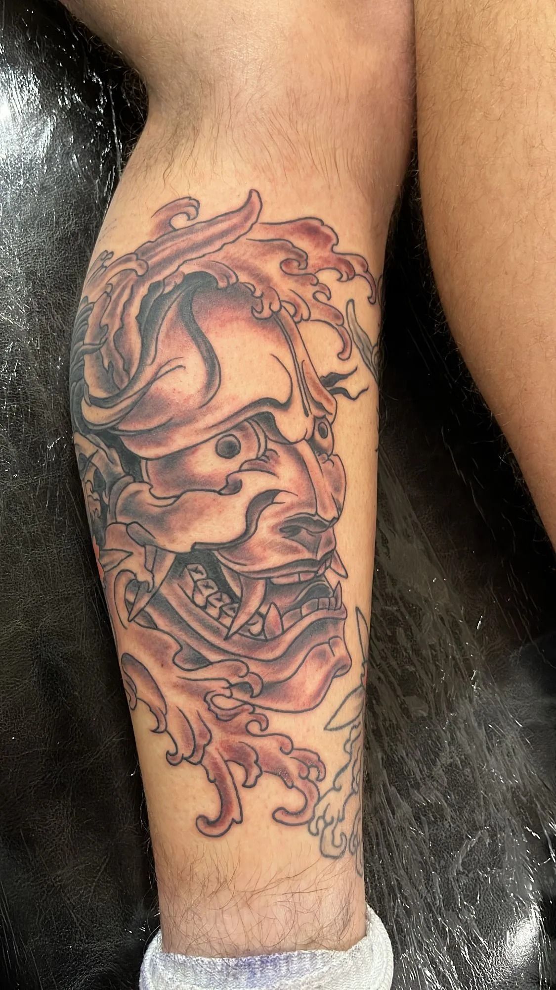 Tattoo of a Japanese demon mask on a leg, rendered in shades of gray, red, and brown, with ornate details.