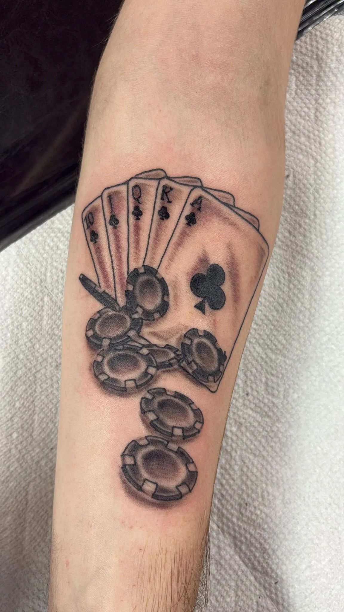 Tattoo of playing cards and poker chips on an arm. Black and gray shading with club suit visible.