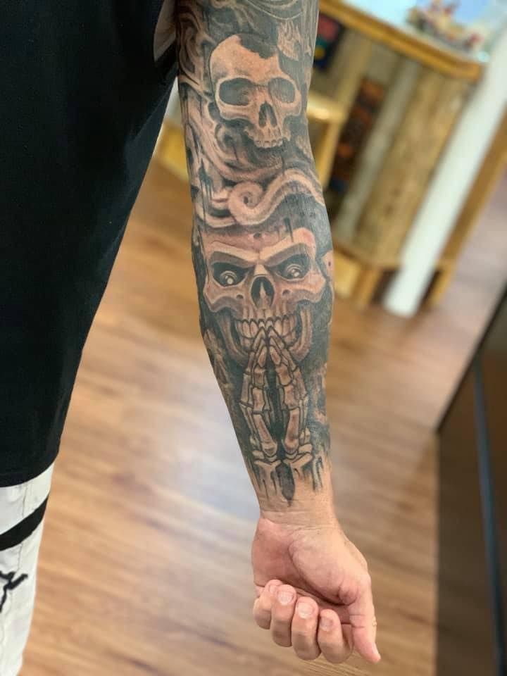 Tattoo of two skulls with smoky swirls on a person's forearm. One skull has clasped skeletal hands in prayer.