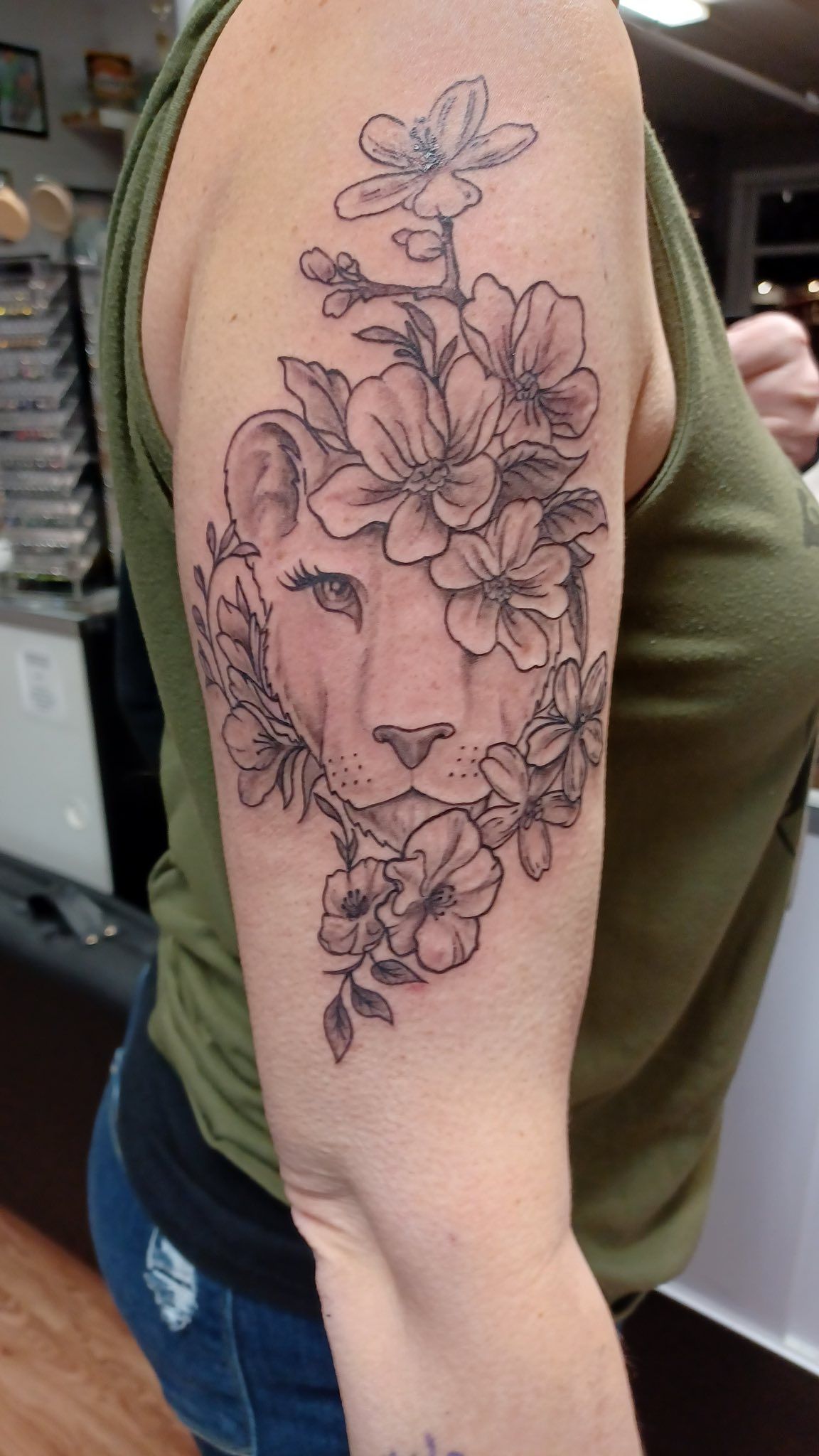 Tattoo of a lioness face with floral accents on a person's upper arm. Black and white ink.