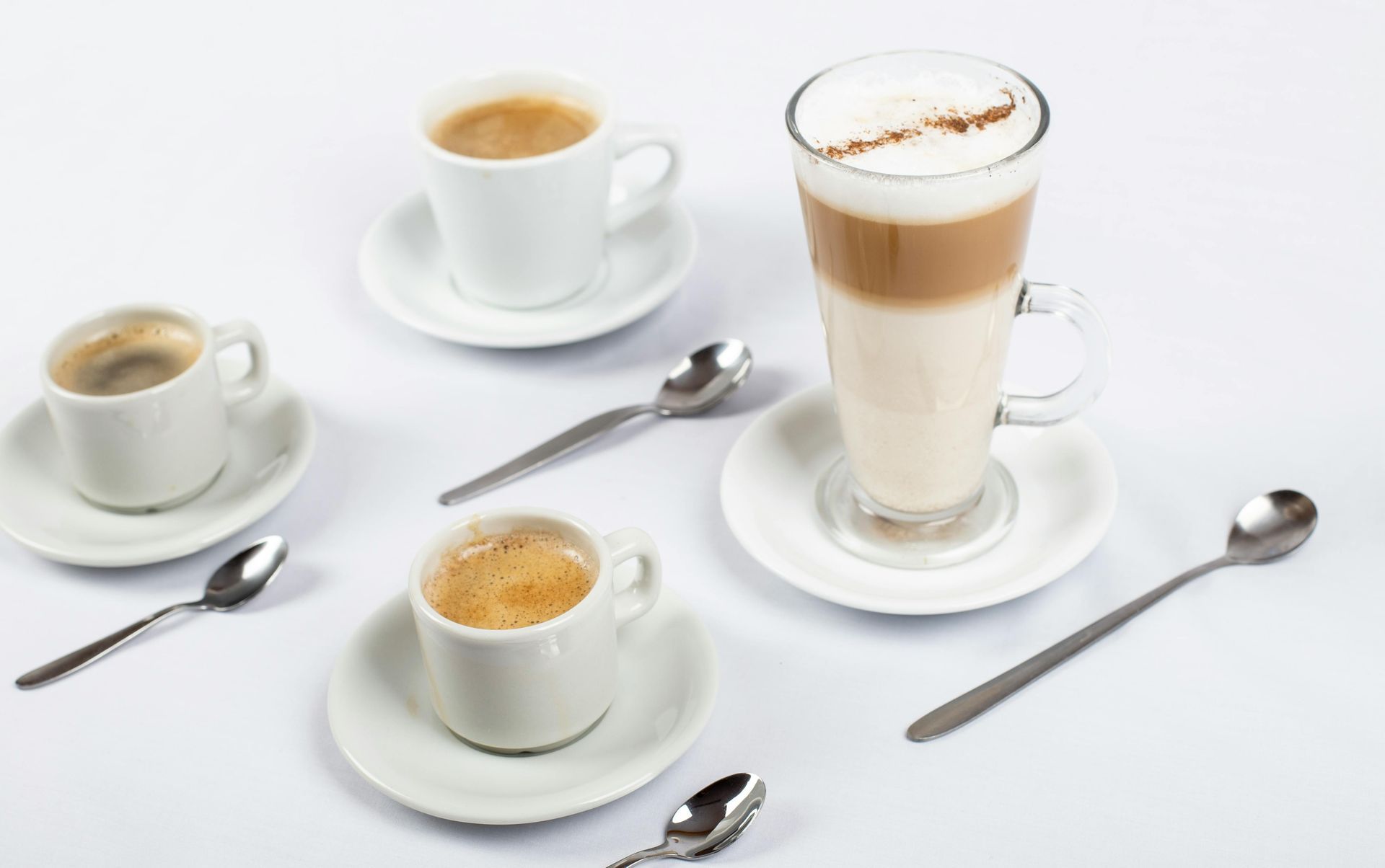 Three small white coffee cups and one tall glass of layered coffee on saucers, arranged with four silver spoons.