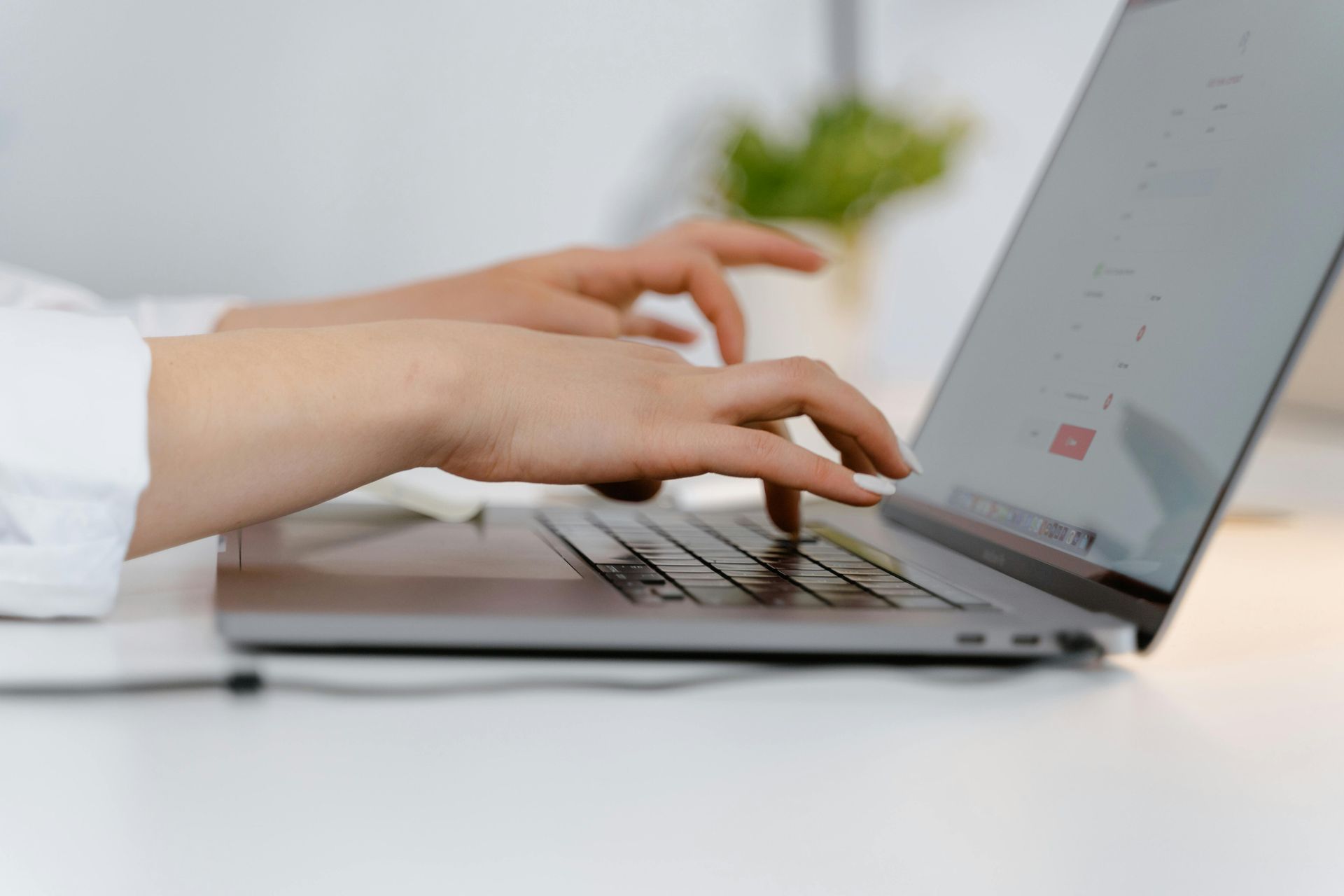 Close-up side view of hands typing on a laptop on a white desk with a blurred green plant in the background.