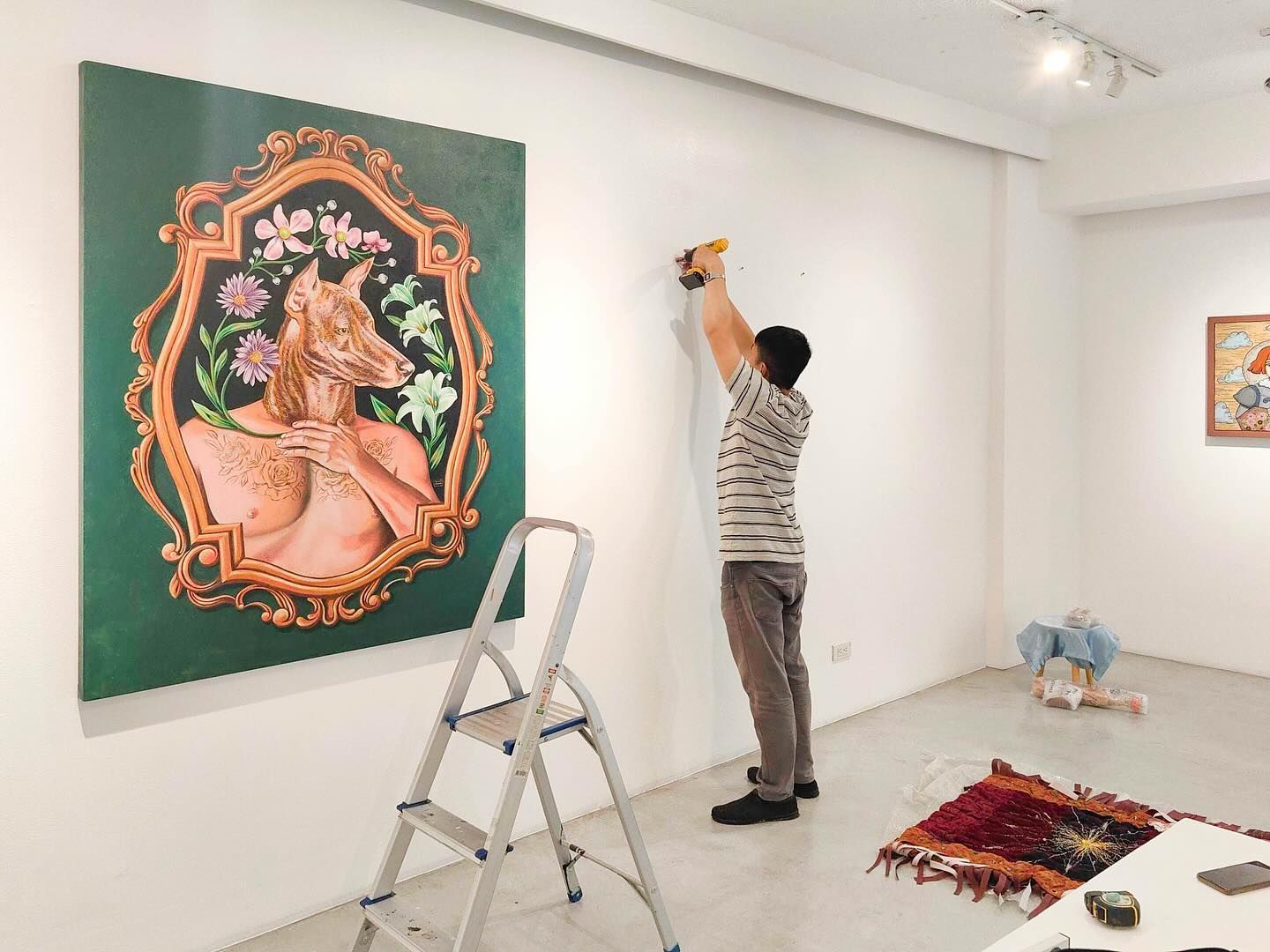 Man on stepladder drilling into a gallery wall, near a framed painting with a fox figure and flowers.
