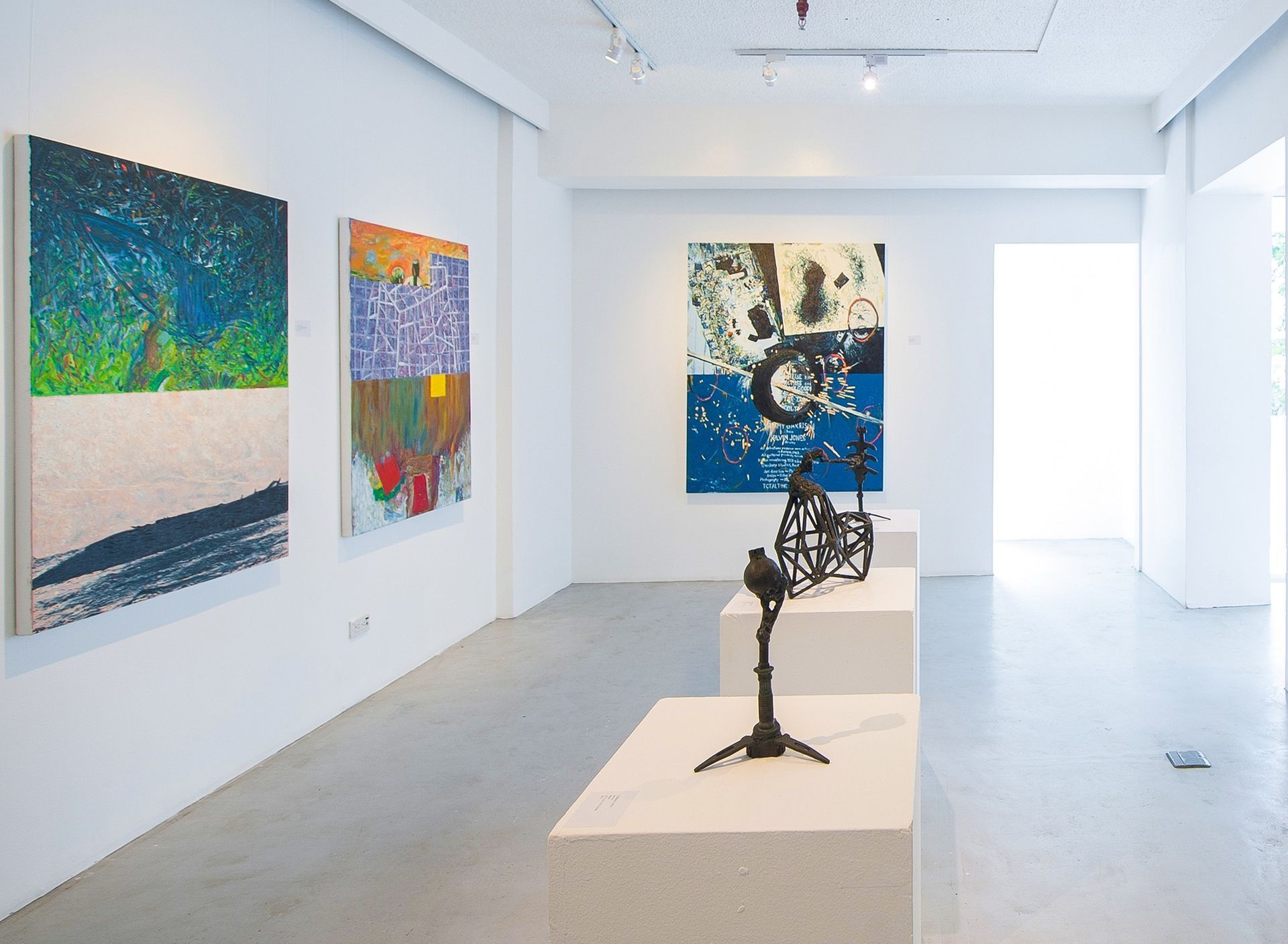 Art gallery with colorful abstract paintings and sculptures on display. Bright white walls and concrete floor.
