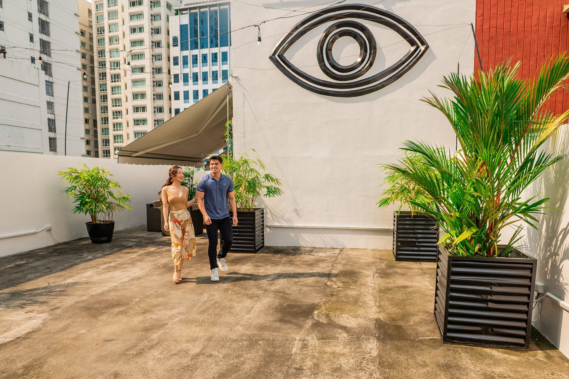Couple walking on rooftop patio with large eye art and potted plants.
