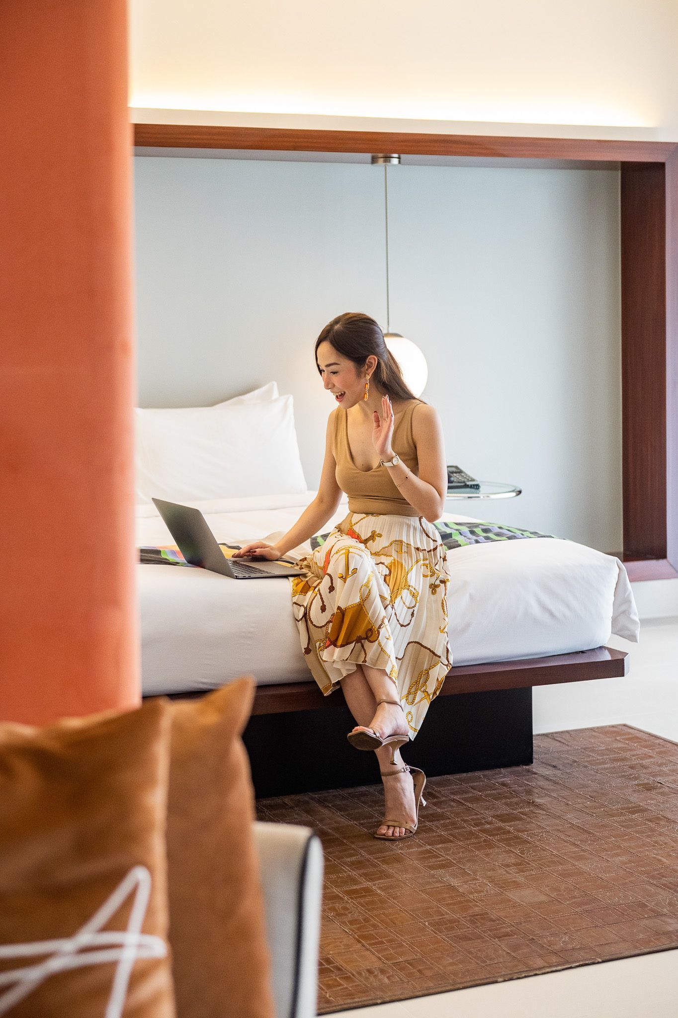 Woman on bed with laptop, waving. In a hotel room with warm color accents.