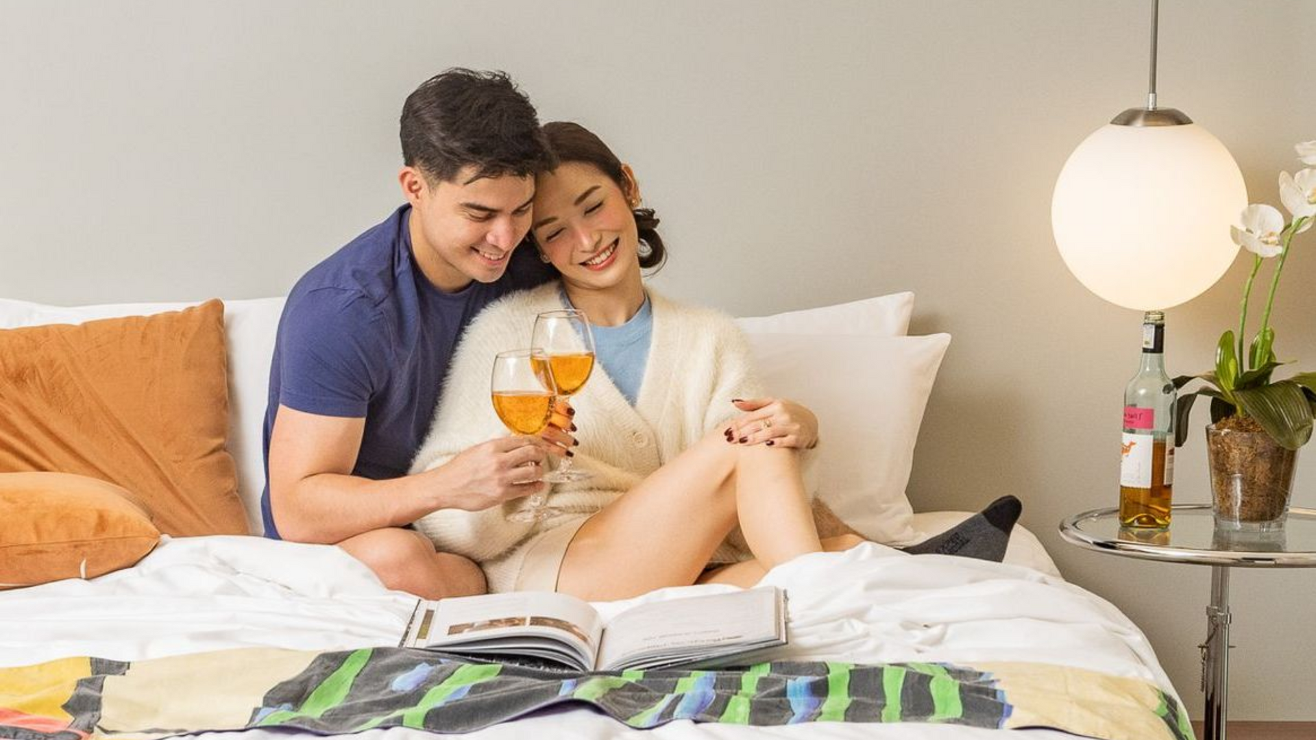 Couple toasting wine on a bed with a book, smiling. Warm tones, modern bedroom setting.