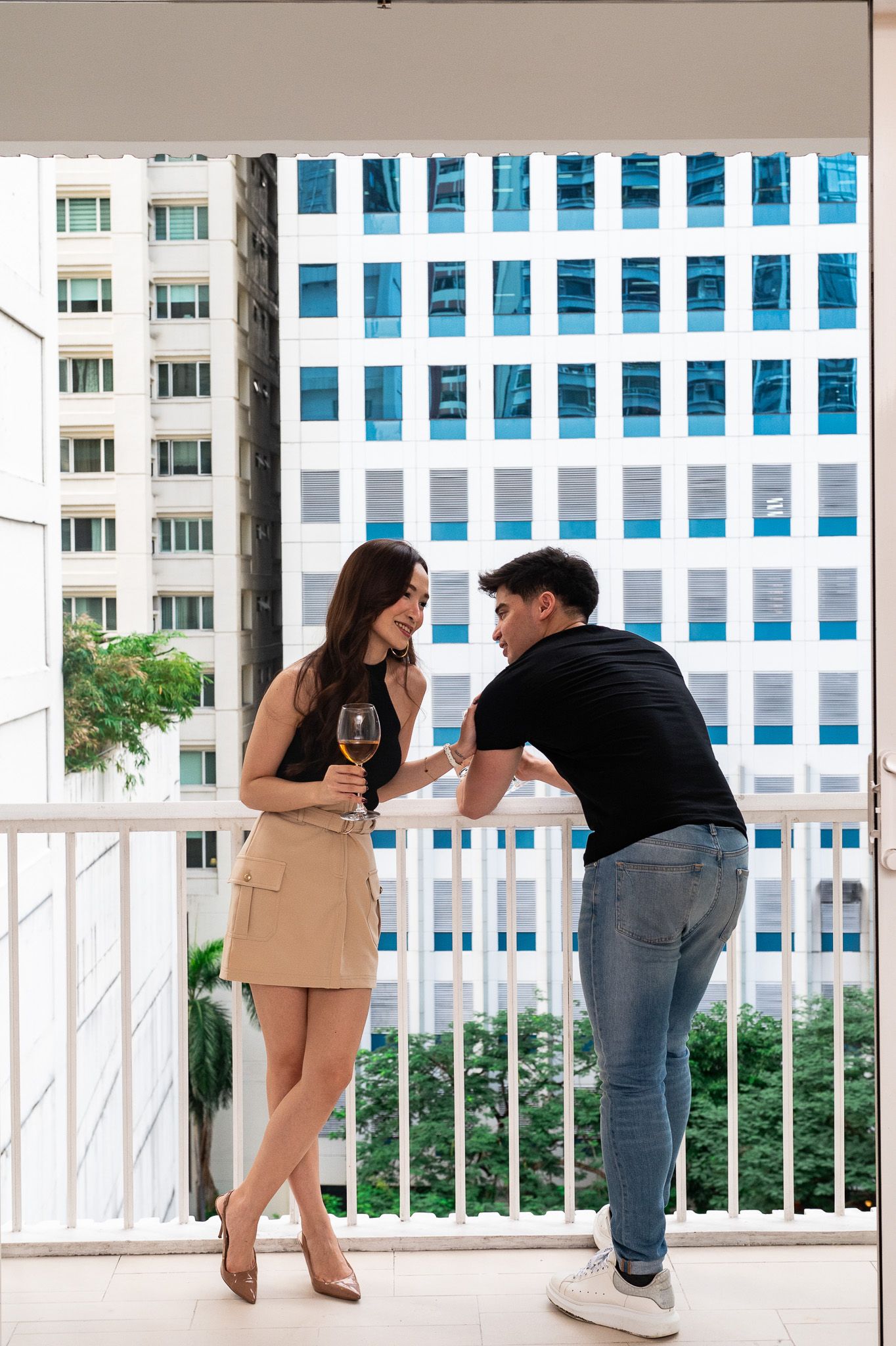 A person in a beige skirt and black top holds a wine glass while talking to a person in a black t-shirt on a balcony.