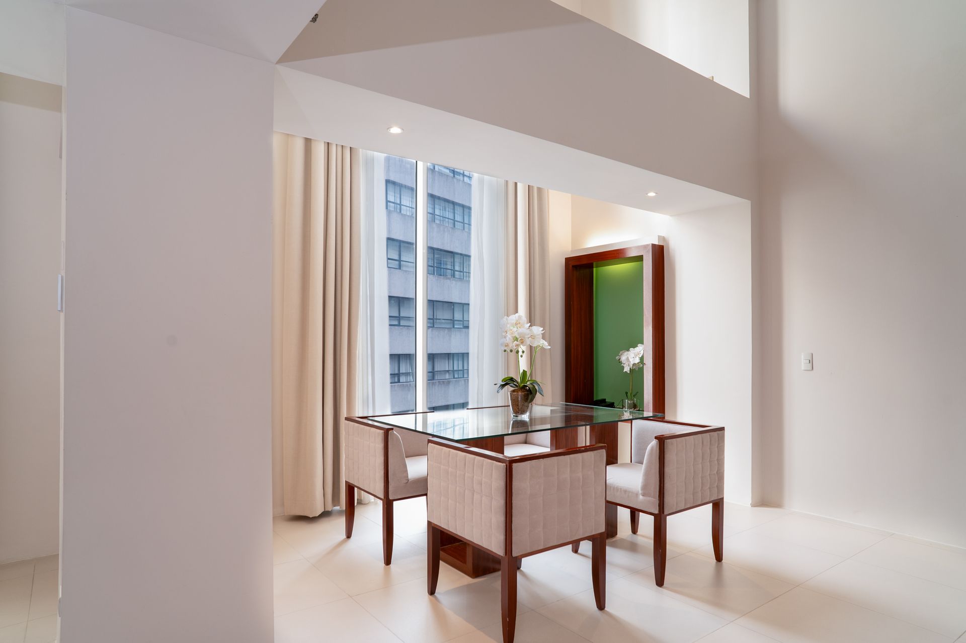 Dining area with a glass-top table, four chairs, and a tall window with neutral curtains; city view.