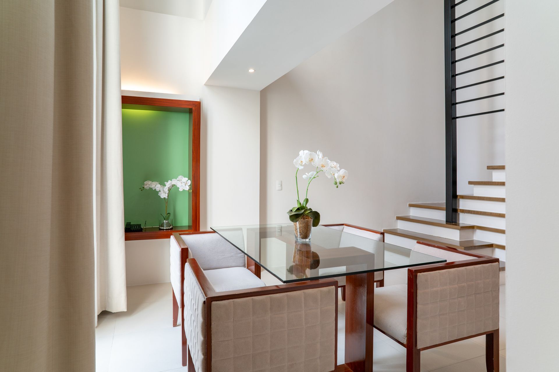 Dining room with glass table, four chairs, orchids, stairs, and a green-framed window.