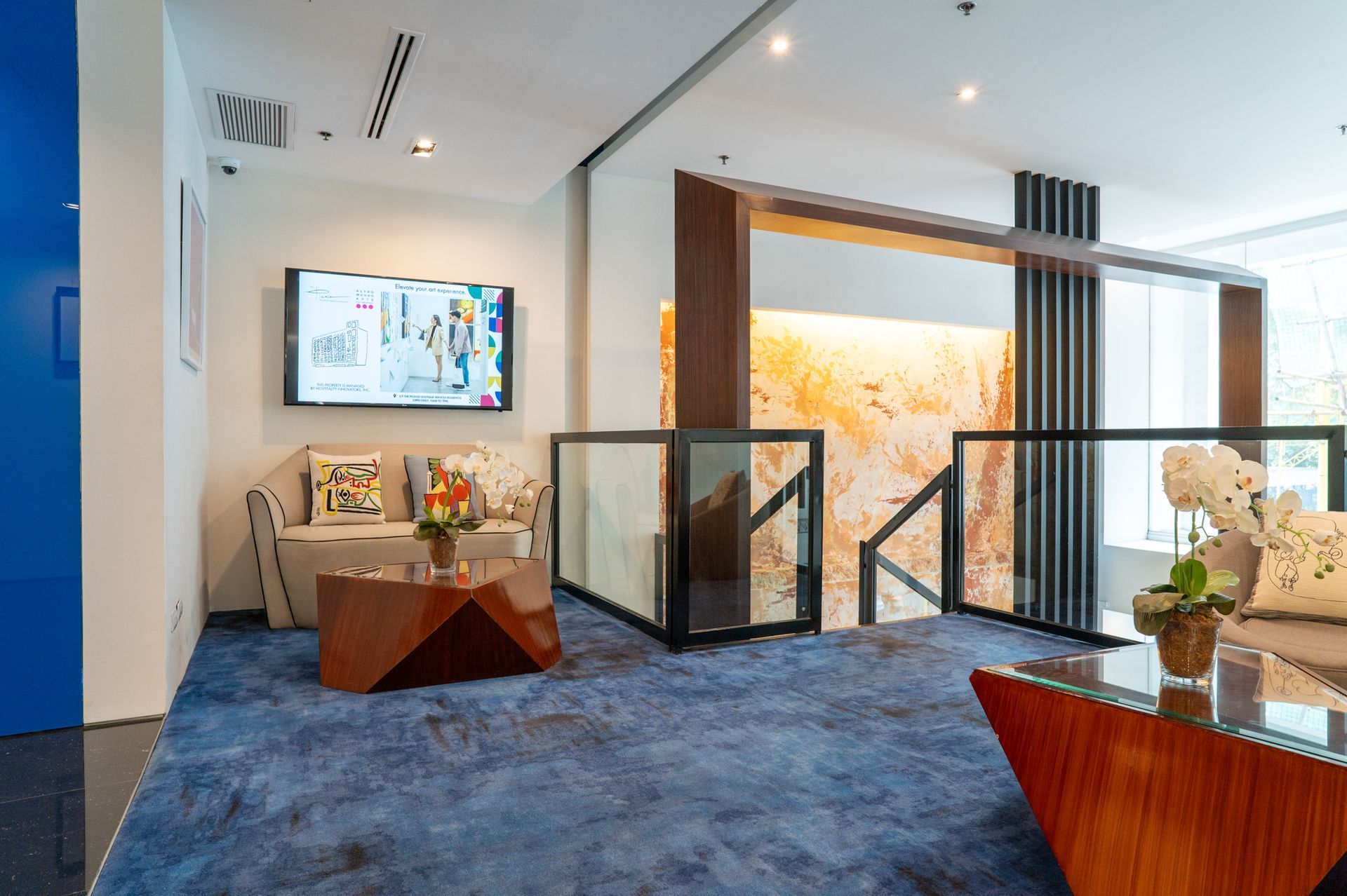 Lobby with blue carpet, couches, TV, and staircase with glass railings and wood accents.