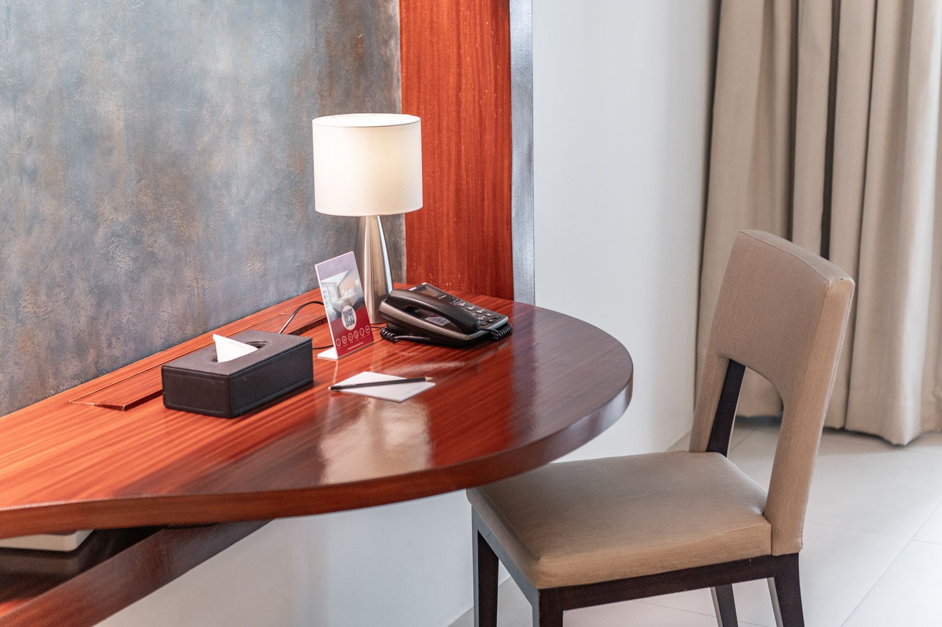 A curved wooden desk with lamp, tissues, and a chair in a room with a painting.