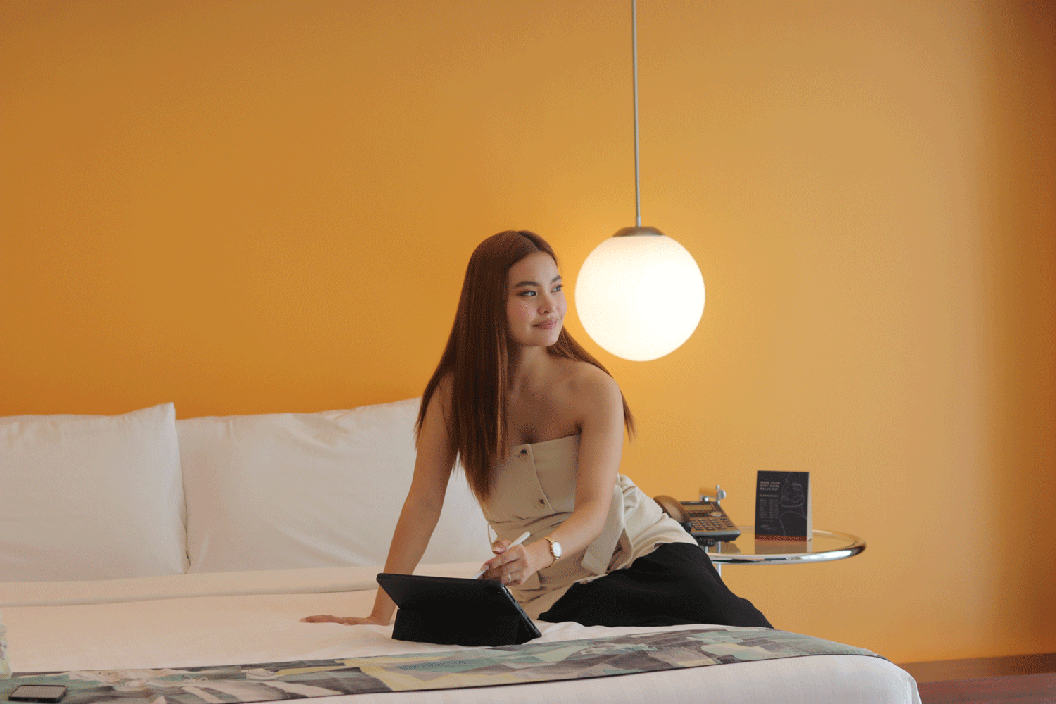 A person sits on a bed in an orange-walled room, using a tablet with a stylus while looking away.