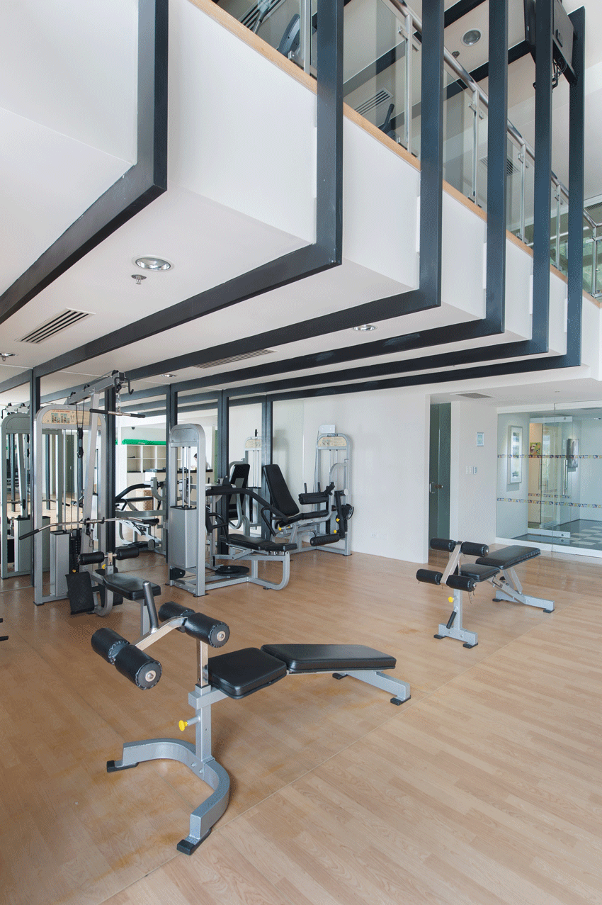 Gym interior with exercise machines and wooden floor. Overhead beams, glass railings, and white walls.