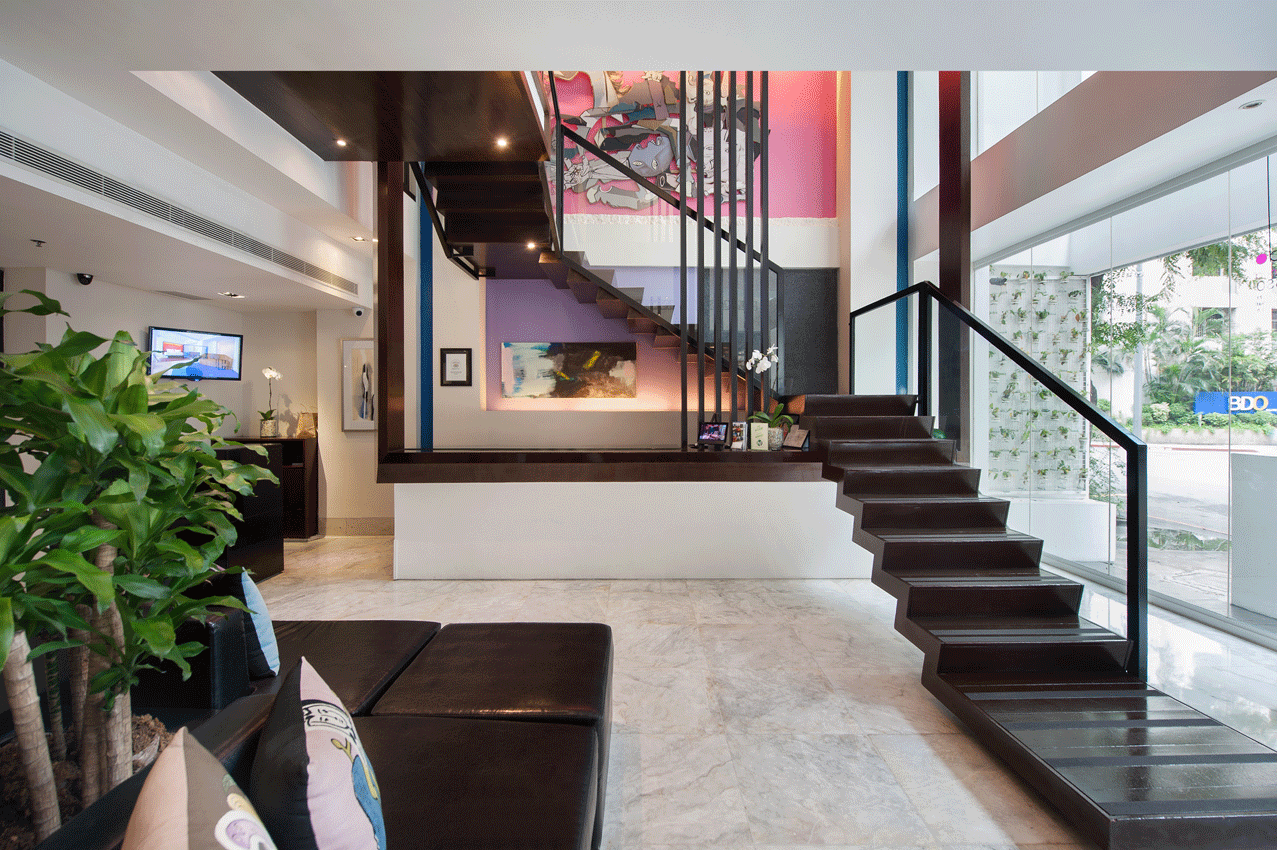 Lobby with staircase leading to upper level. Black sofa, marble floor, large art piece, plants, and glass windows.