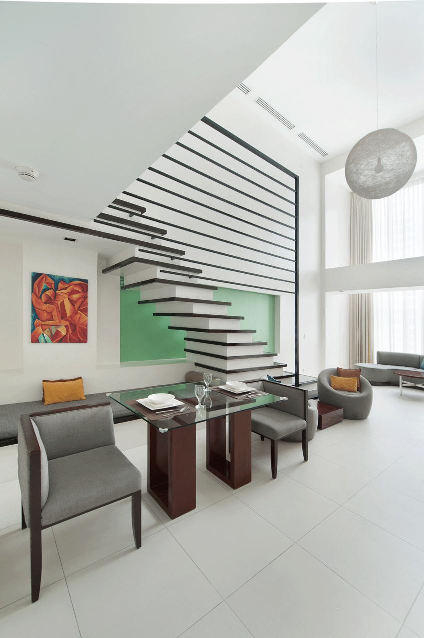 A modern living space features a glass dining table, grey chairs, and a floating staircase against a green wall.