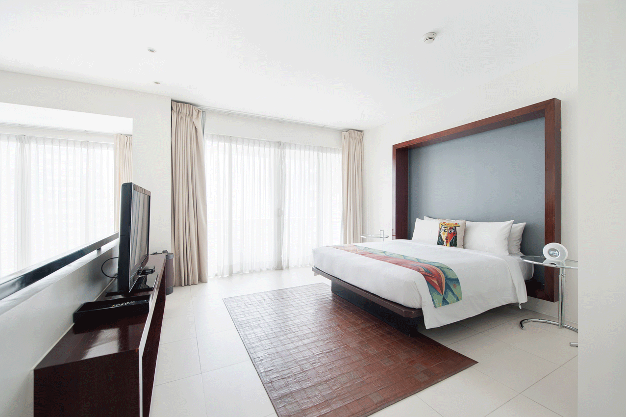 A modern hotel bedroom with a large bed, brown area rug, floor-to-ceiling windows with curtains, and a flat-screen TV.