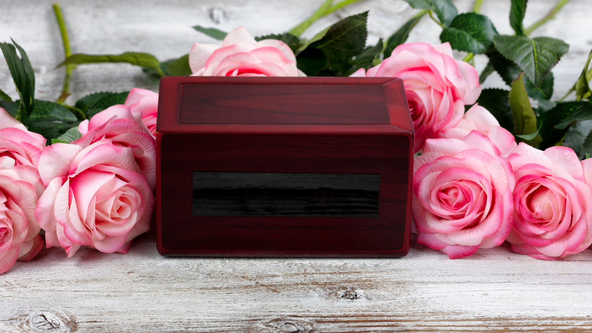 An image of a brown cremation box sitting on a white faded wooden table with pink roses beside it