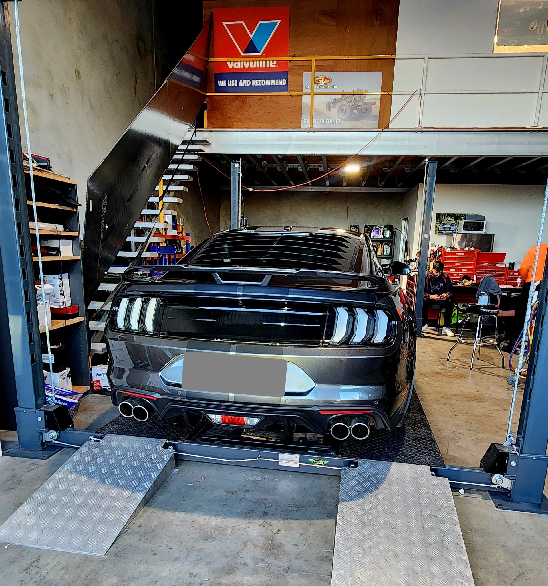 Dark Gray Sports Car on a Lift in a Garage. Shop Sign and Stairs Visible in Background — QA Oxley in Sherwood, QLD