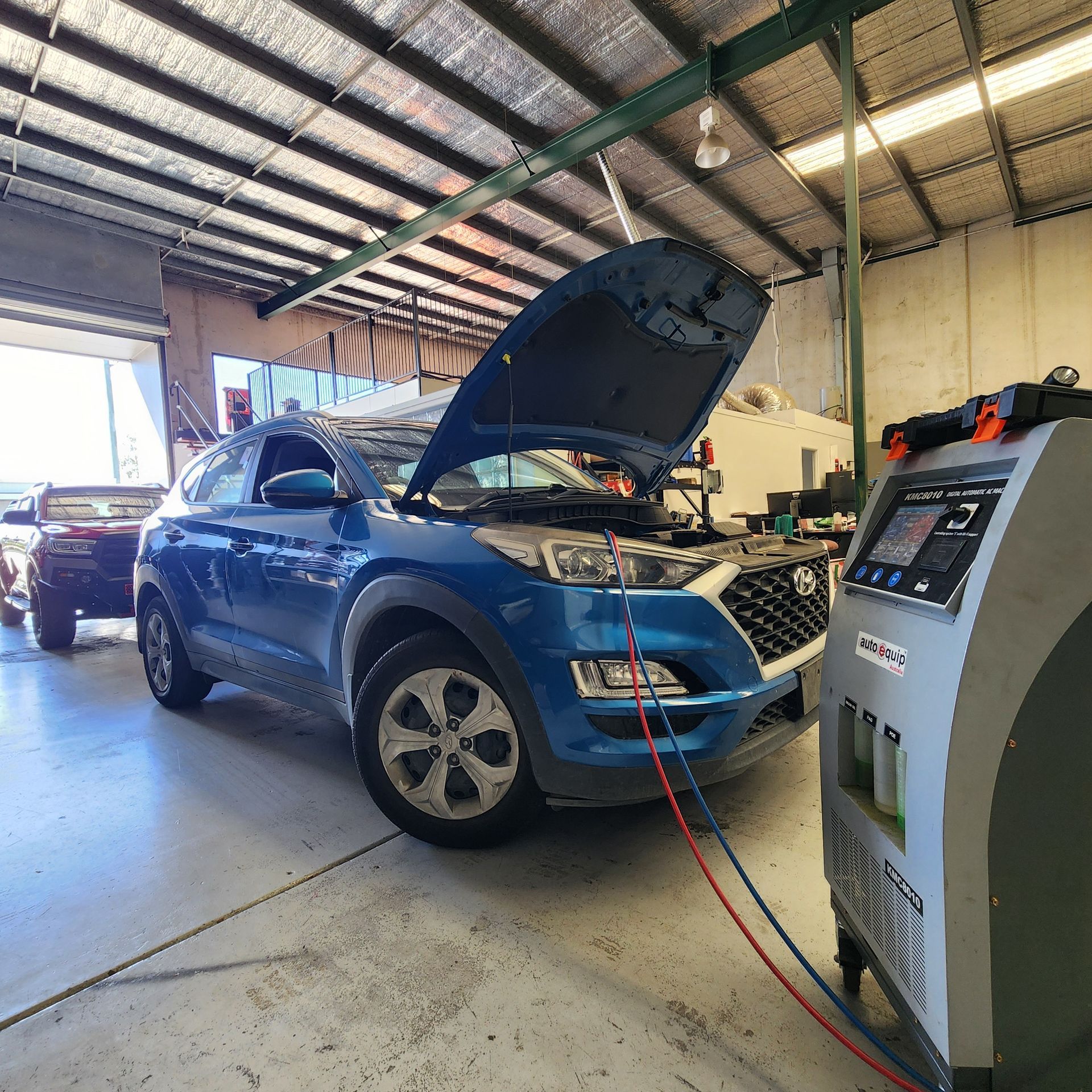 A blue car in a garage with the hood open, connected to an air conditioning service machine.